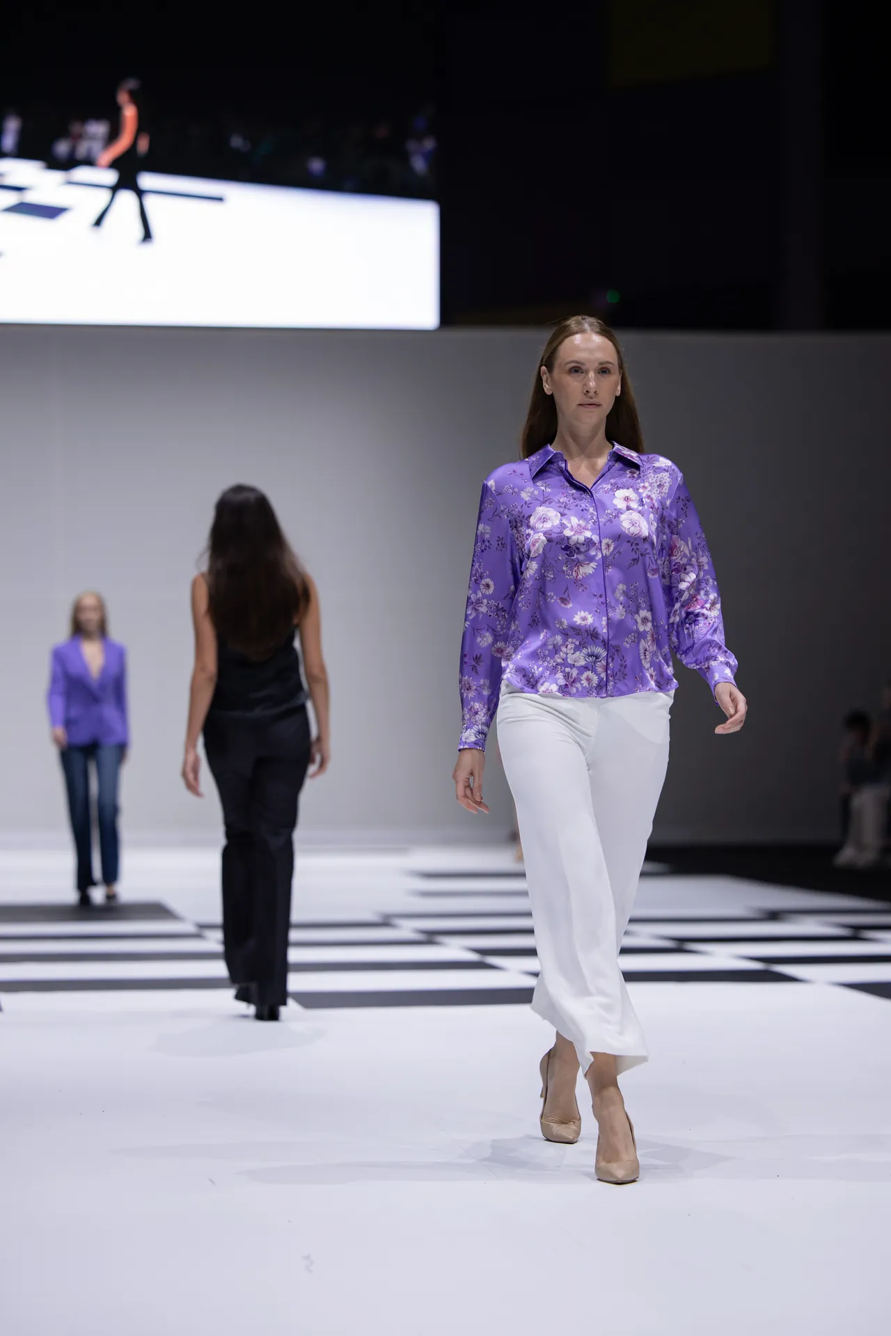 Model walks down a white and black checkered runway in a purple floral blouse and white pants, others in background wearing similar outfits