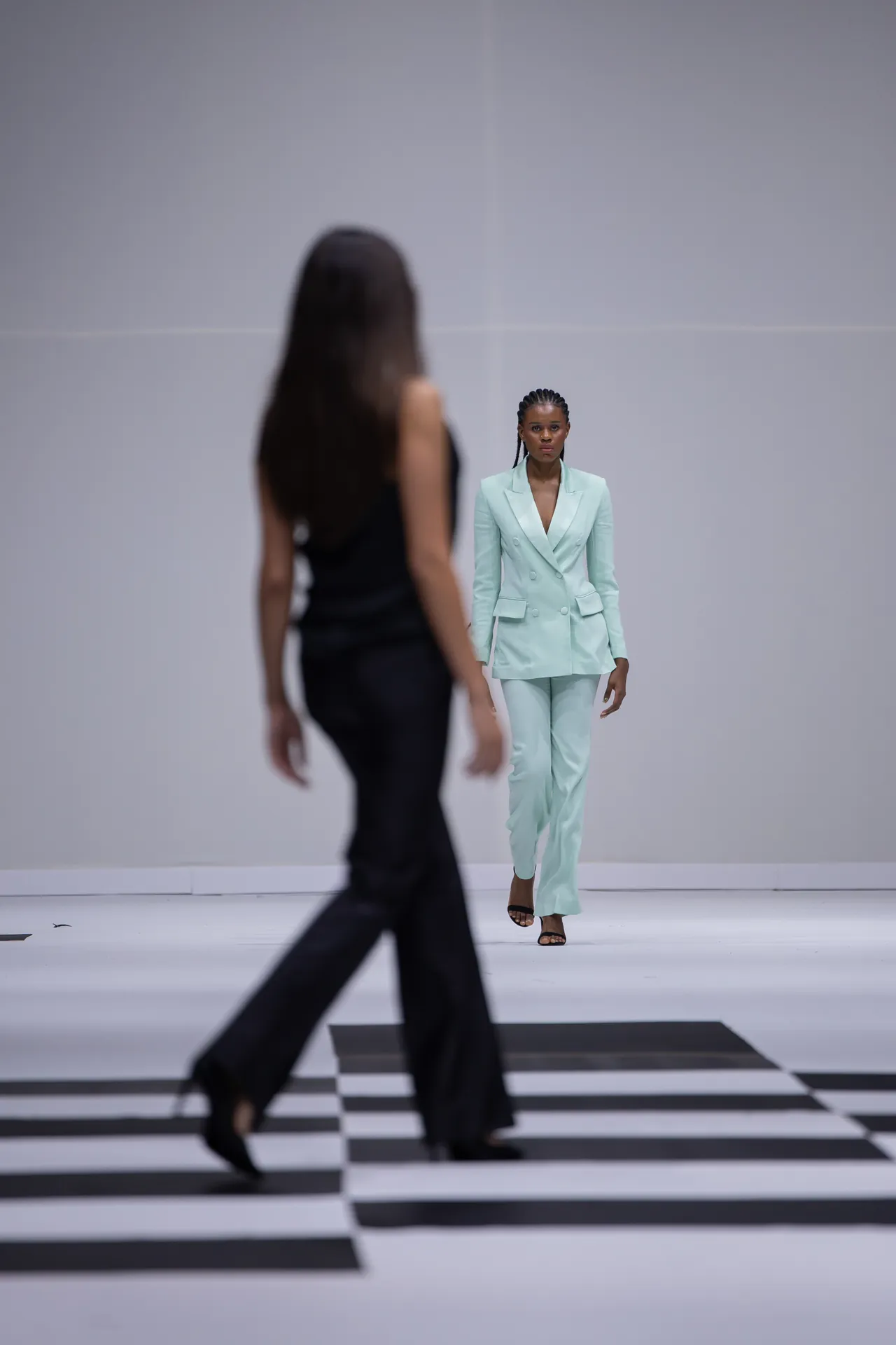 Model in a mint green double-breasted suit walks a black-and-white striped runway; foreground silhouette of another model blur.