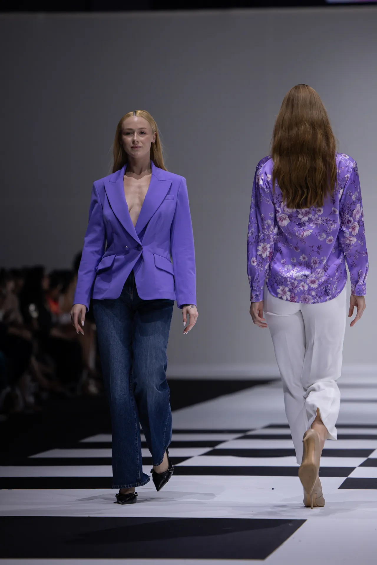 Two models walk a runway in purple outfits: one in a lavender blazer with jeans, the other in a purple floral blouse and white pants (back view).