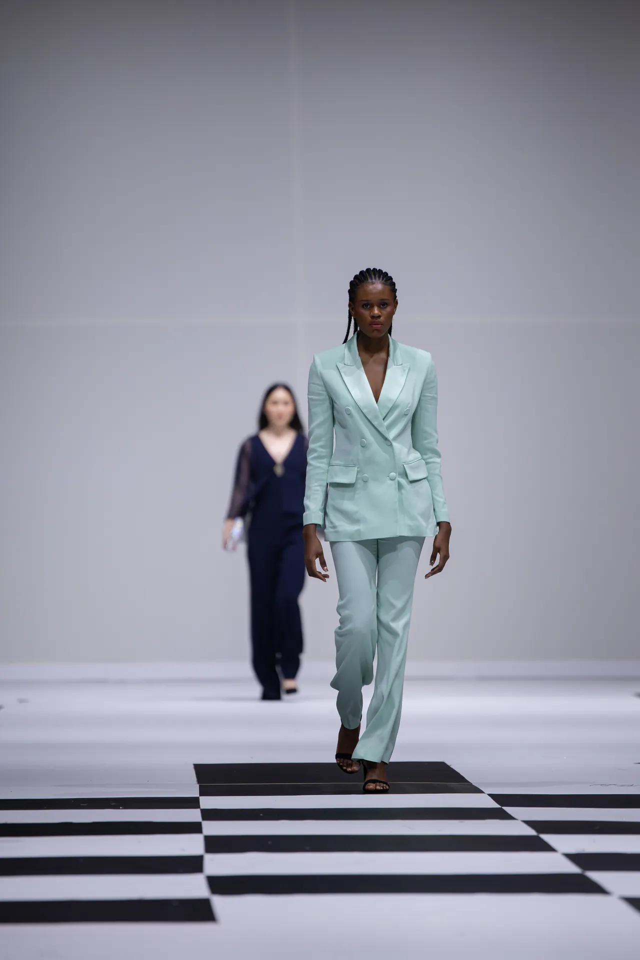 Female model walks a mint-green tailored pantsuit down a black-and-white checkered runway, focused and poised.