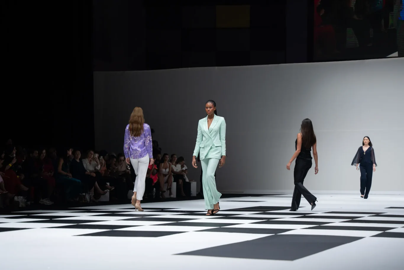 Model in a mint green suit walks down a black-and-white geometric runway as the audience watches from the left side of the catwalk.