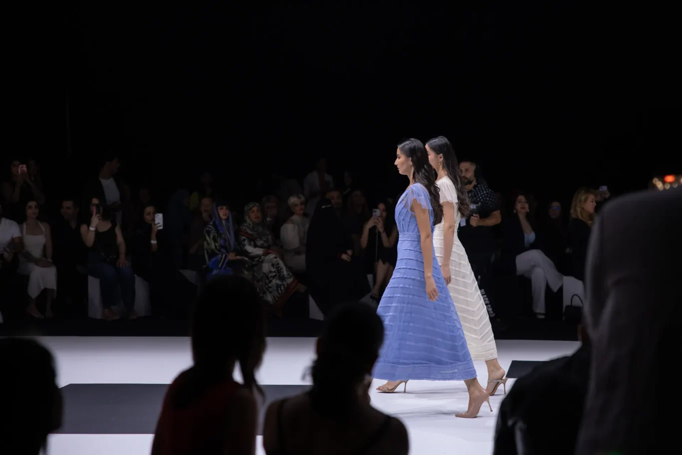 Two female models walk a fashion runway in blue and white striped dresses as an audience watches from darkened seats nearby.
