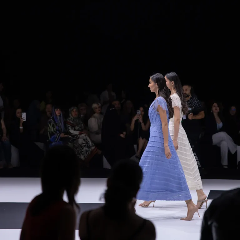 Two female models walk a fashion runway in blue and white striped dresses as an audience watches from darkened seats nearby.