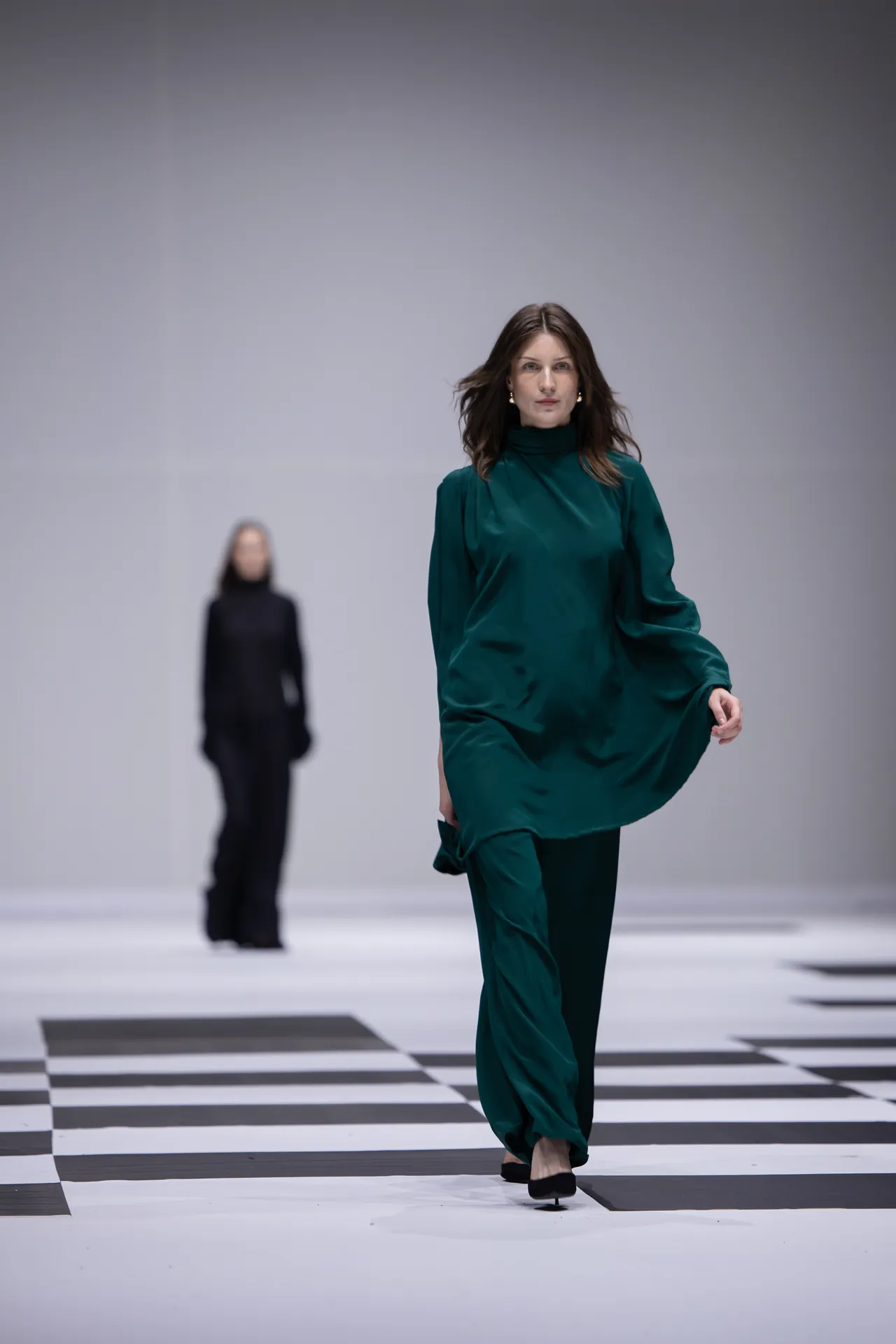 Model in emerald green outfit walks a black-and-white checkered runway; another model in black blurred in the background.