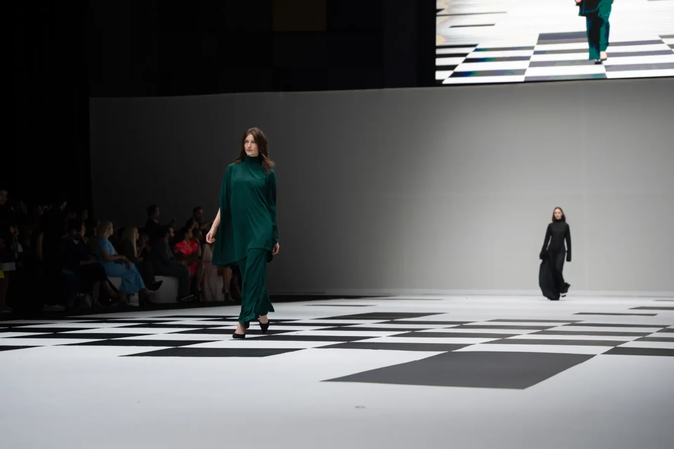 Model walks down a black-and-white checkered runway in a green outfit as spectators watch from the left.