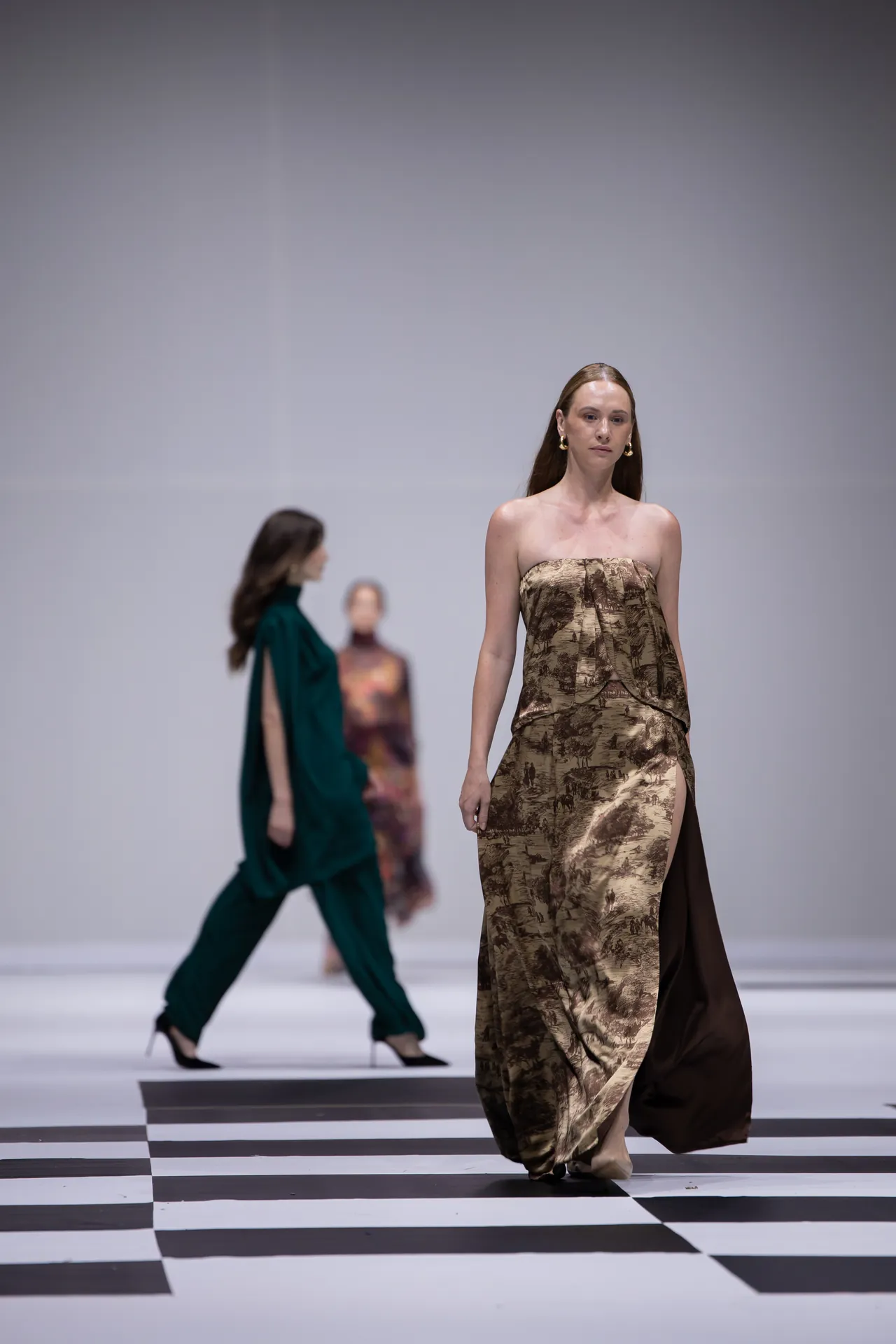 Model walks a fashion show wearing a strapless bronze-brown patterned gown on a black-and-white checkered runway.