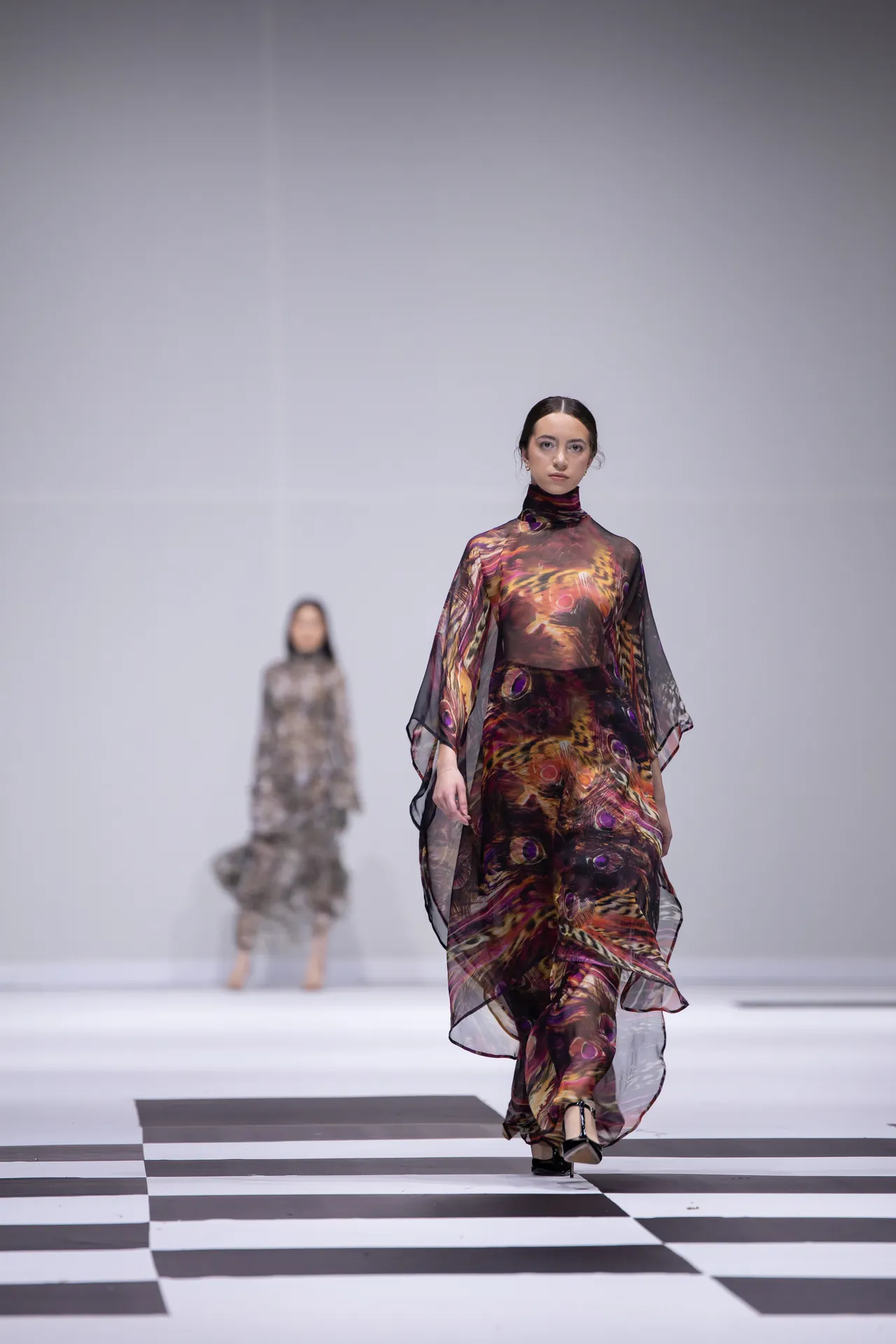 Model walks a runway in a flowing, multicolored sheer gown; a checkered floor and a blurred second model in the background.