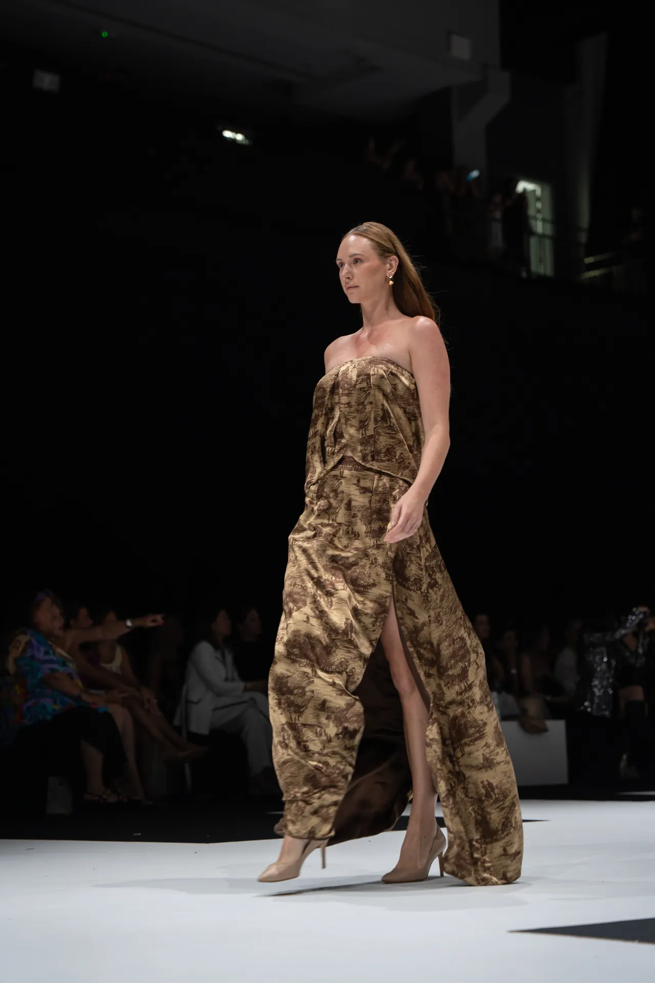 Model walking a runway in a brown patterned strapless gown with a high slit, beige heels, at a fashion show.