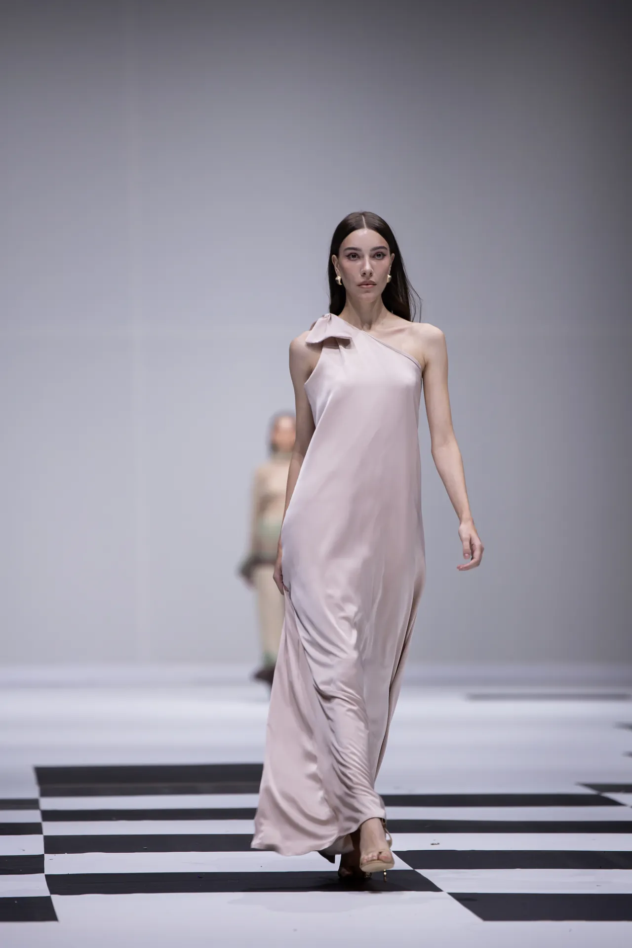 Model walking the runway in a pale pink satin one-shoulder gown on a black-and-white checkered floor, during a fashion show.