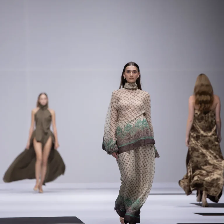 Model walking a fashion runway in a beige patterned outfit with green trim, foreground, other models in background on a black-and-white striped catwalk
