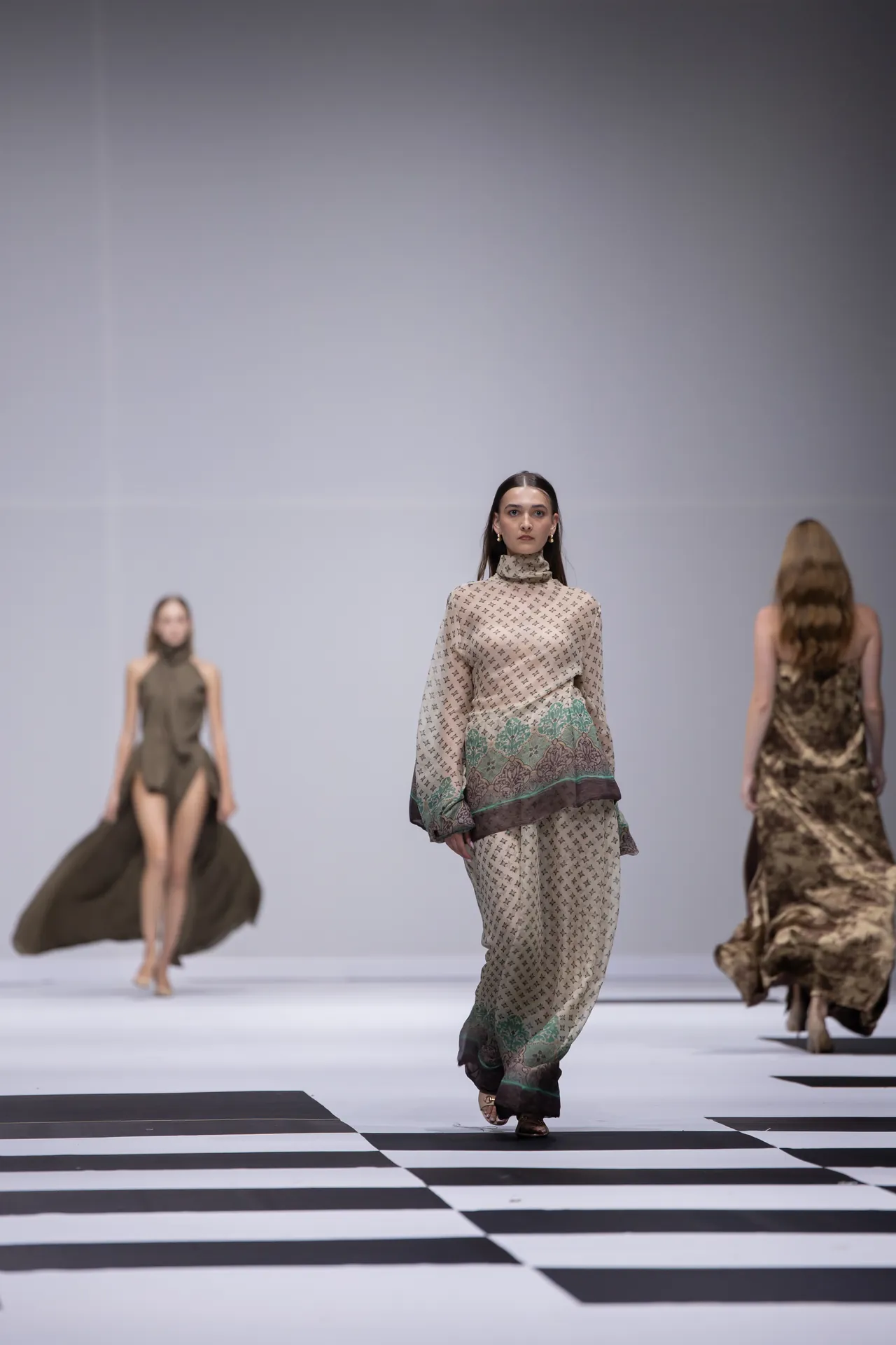 Model walking a fashion runway in a beige patterned outfit with green trim, foreground, other models in background on a black-and-white striped catwalk