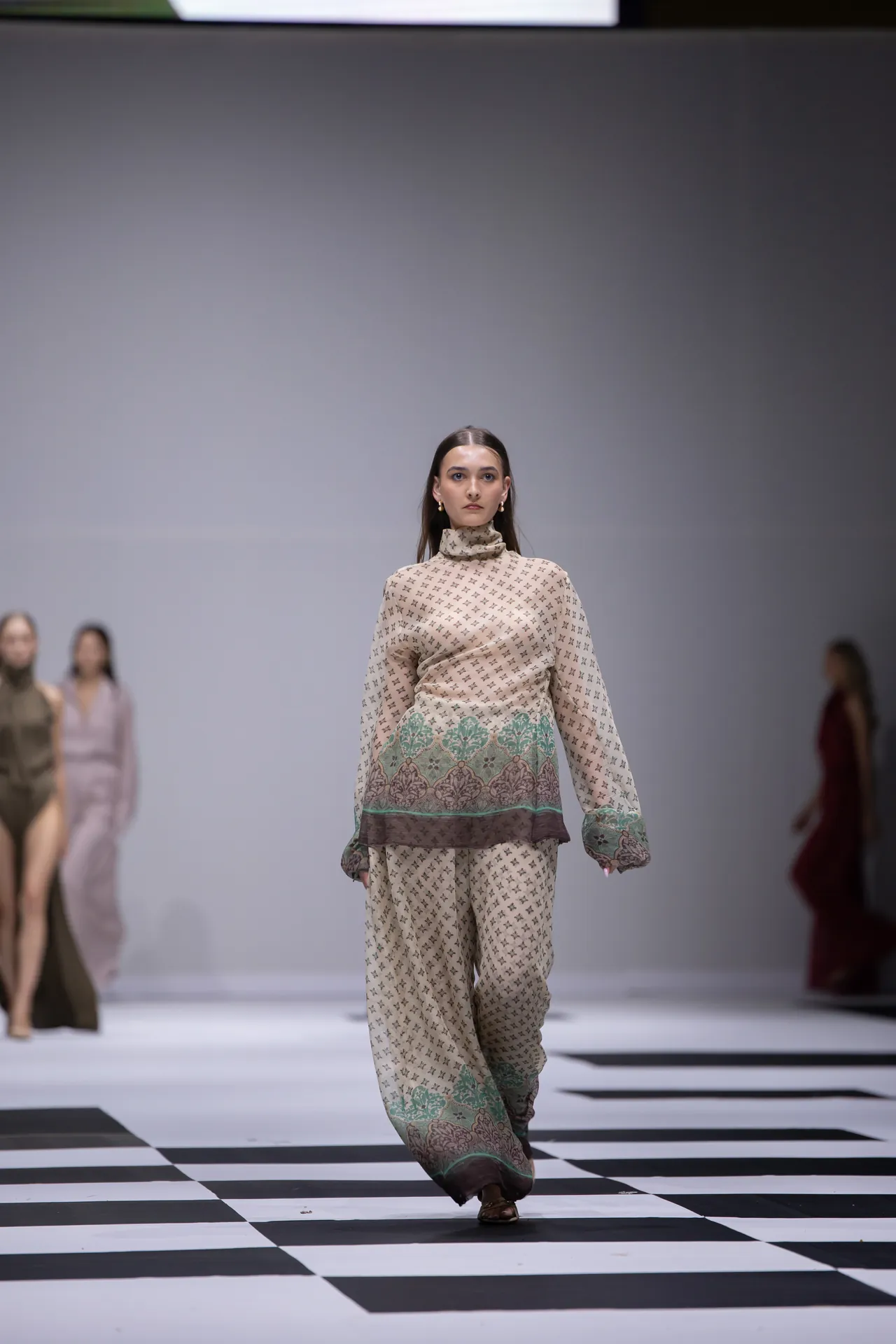 Model walks down a fashion runway in a beige patterned outfit with green embroidery along the hem; black-and-white checkered floor and blurred background models visible in the distance.