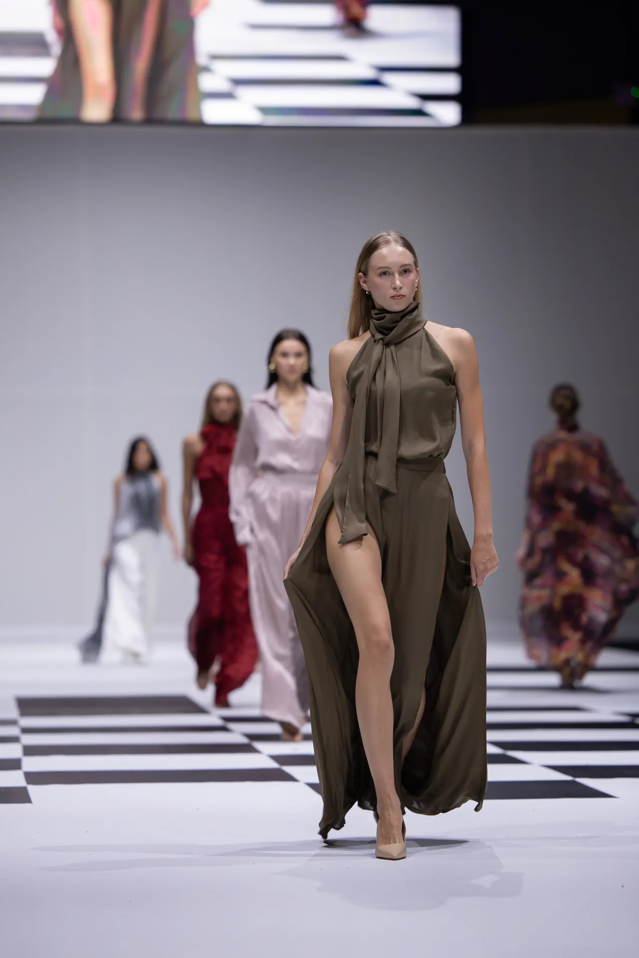 Model in an olive draped halter dress walks a black-and-white checkerboard runway, with other models in colorful outfits behind her.