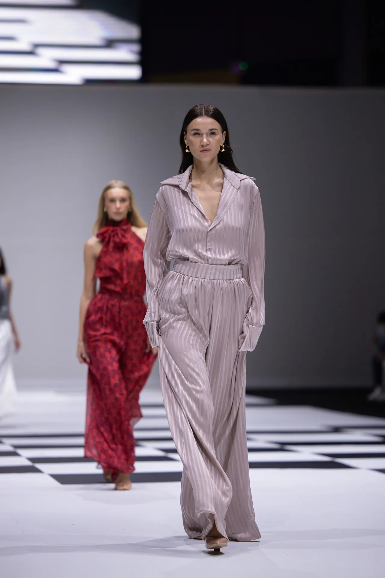 Model walks the runway in a pale pink, pinstriped jumpsuit; another model in red dress behind her on a black-and-white checkered catwalk.