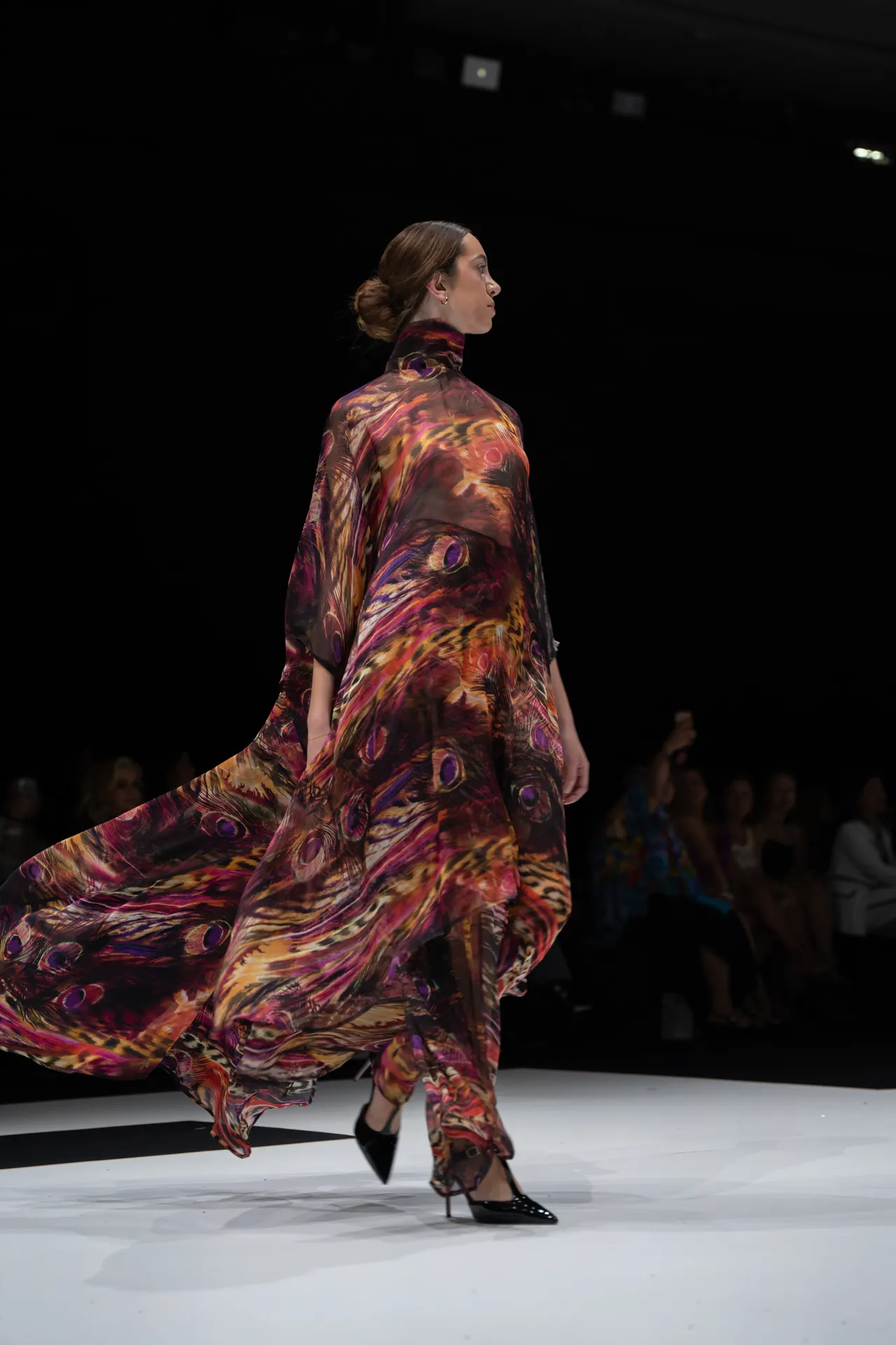 Model walking a fashion runway in a flowing, multicolored print dress; side profile with audience dimly visible in the background.
