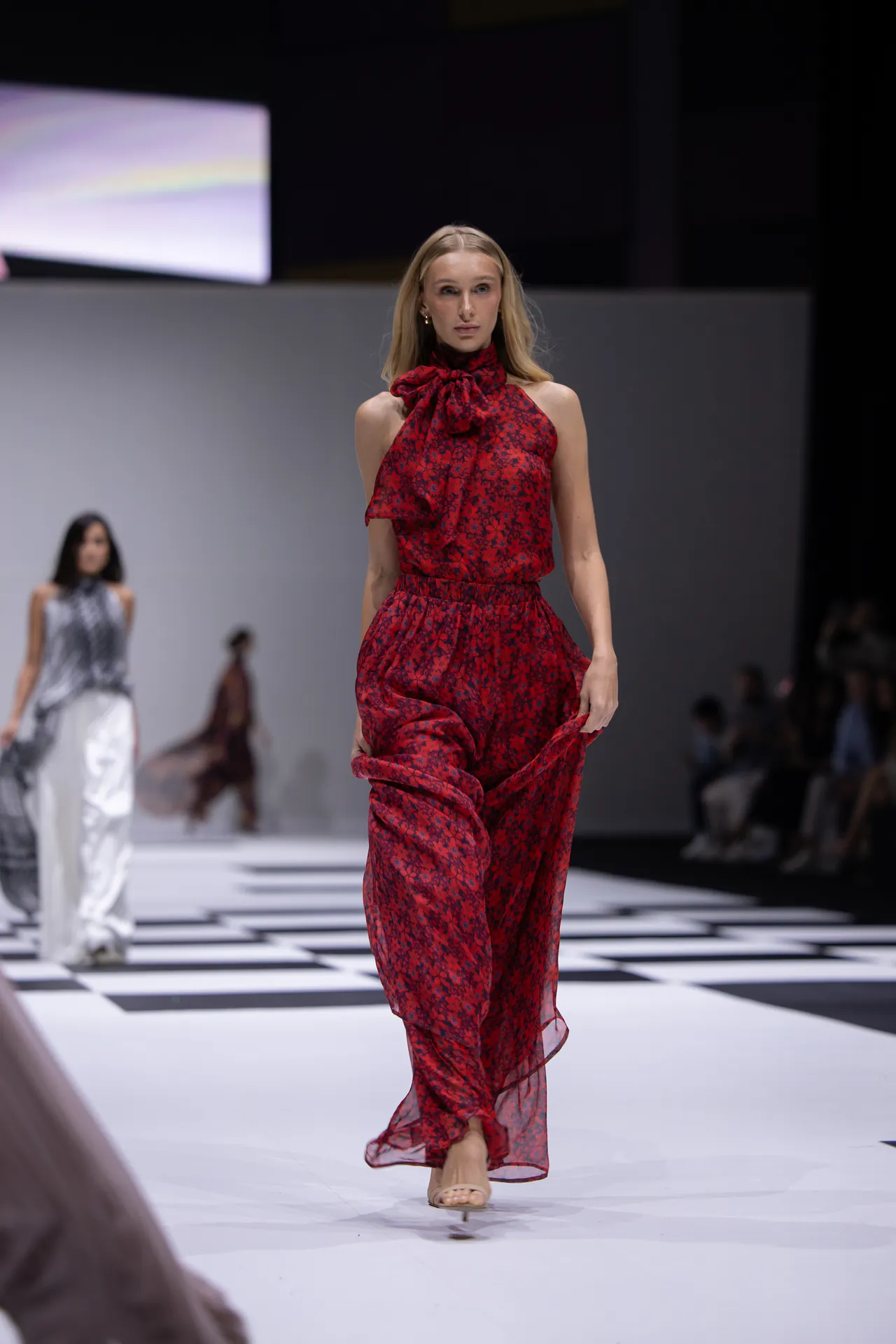 Model walks the fashion runway in a red floral halter jumpsuit with a large bow, on a black-and-white checkered floor with blurred background models.