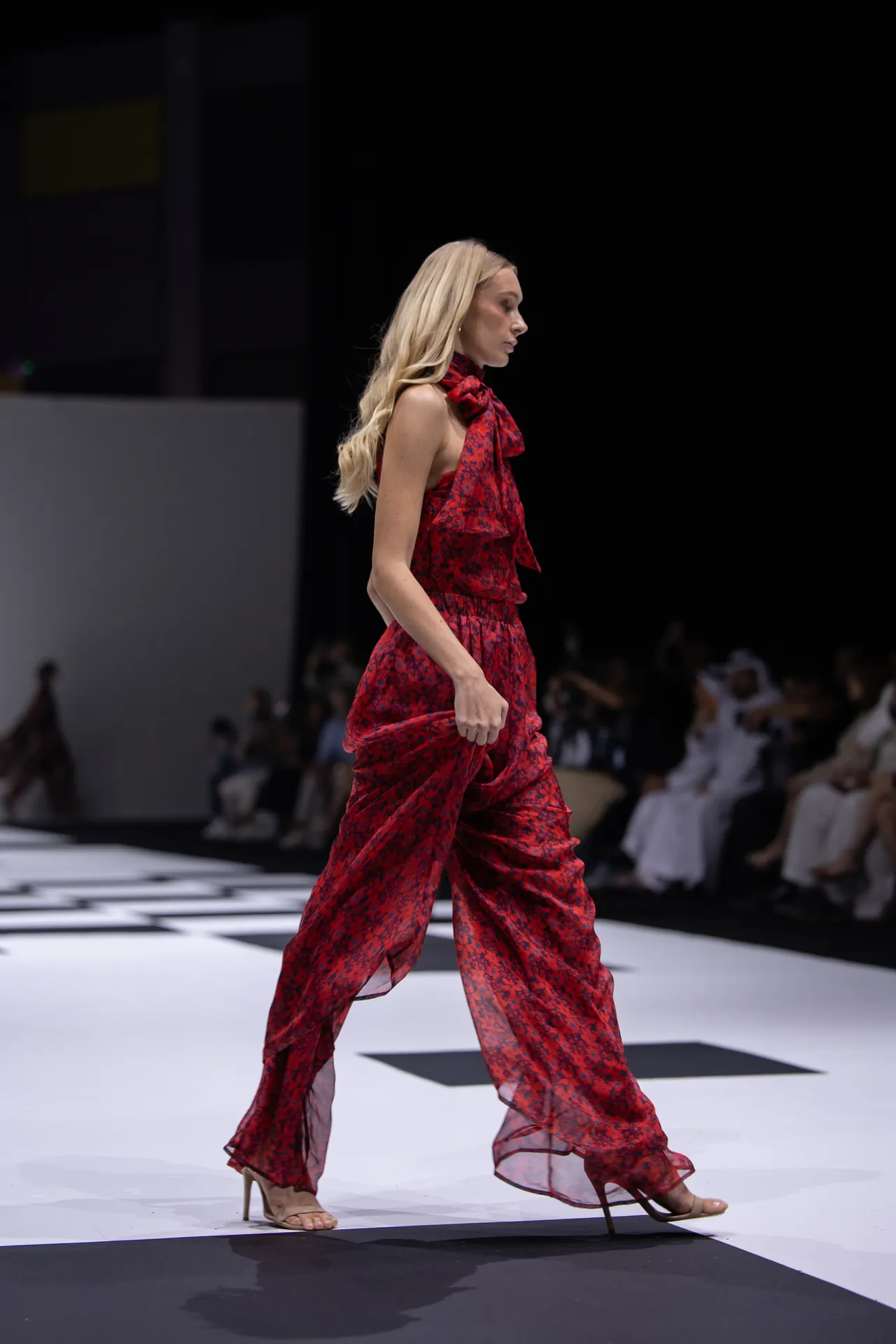 Model walks a red patterned chiffon dress down a white runway, audience in background.