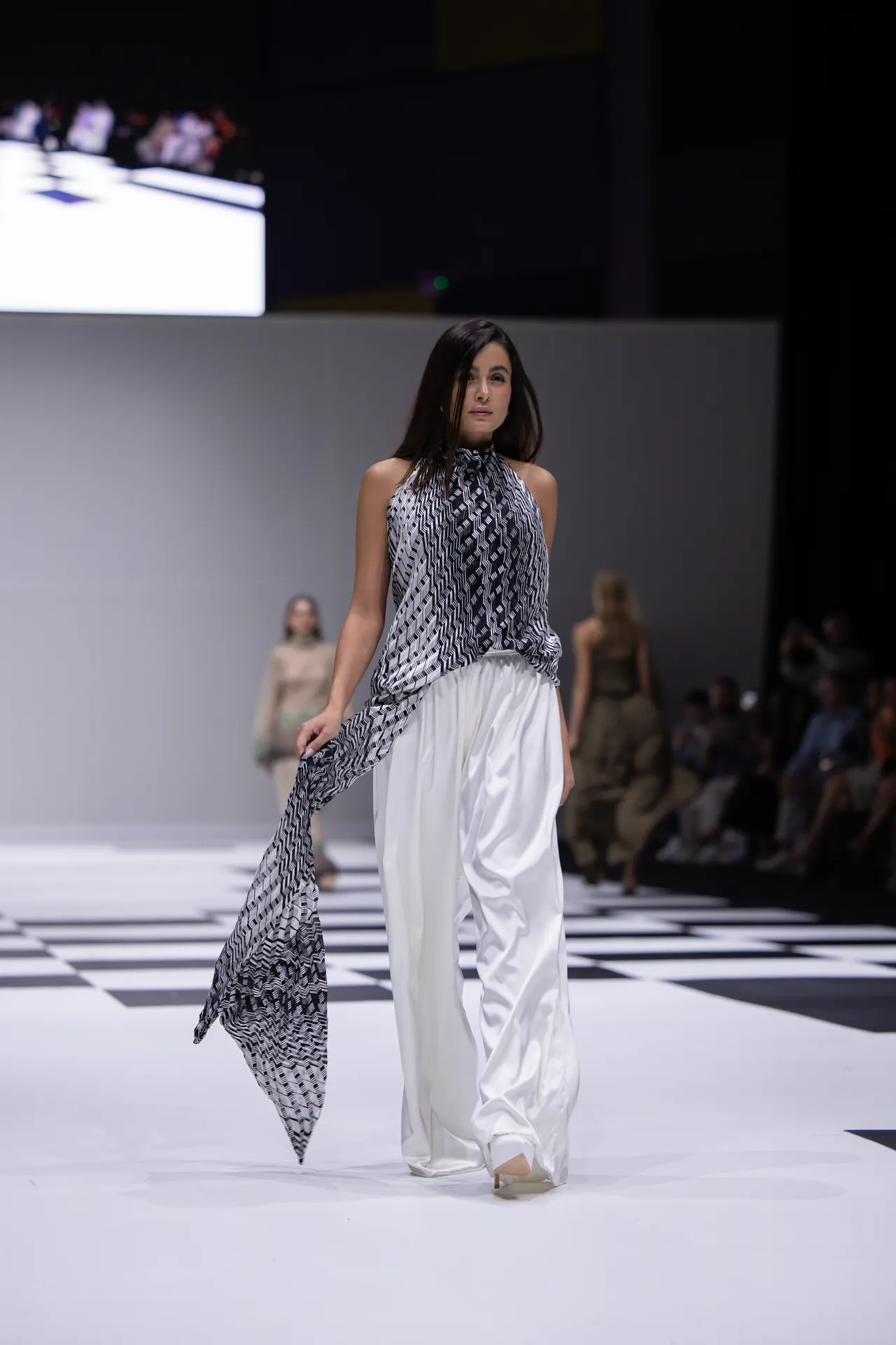 Model walks a runway in a sleeveless black-and-white geometric halter top with flowing white pants; a long matching scarf trails behind her as she strides forward.