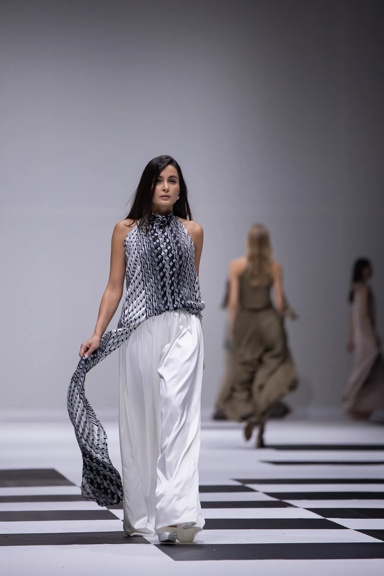 Female fashion model walking down a black-and-white checkerboard runway in a patterned halter top and white wide-leg pants.
