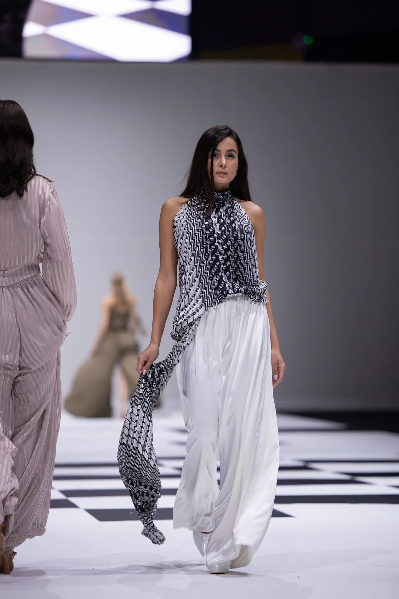 Model walking the runway in a black-and-white geometric halter top and flowing white pants on a checkered catwalk while another designer garment is visible nearby.