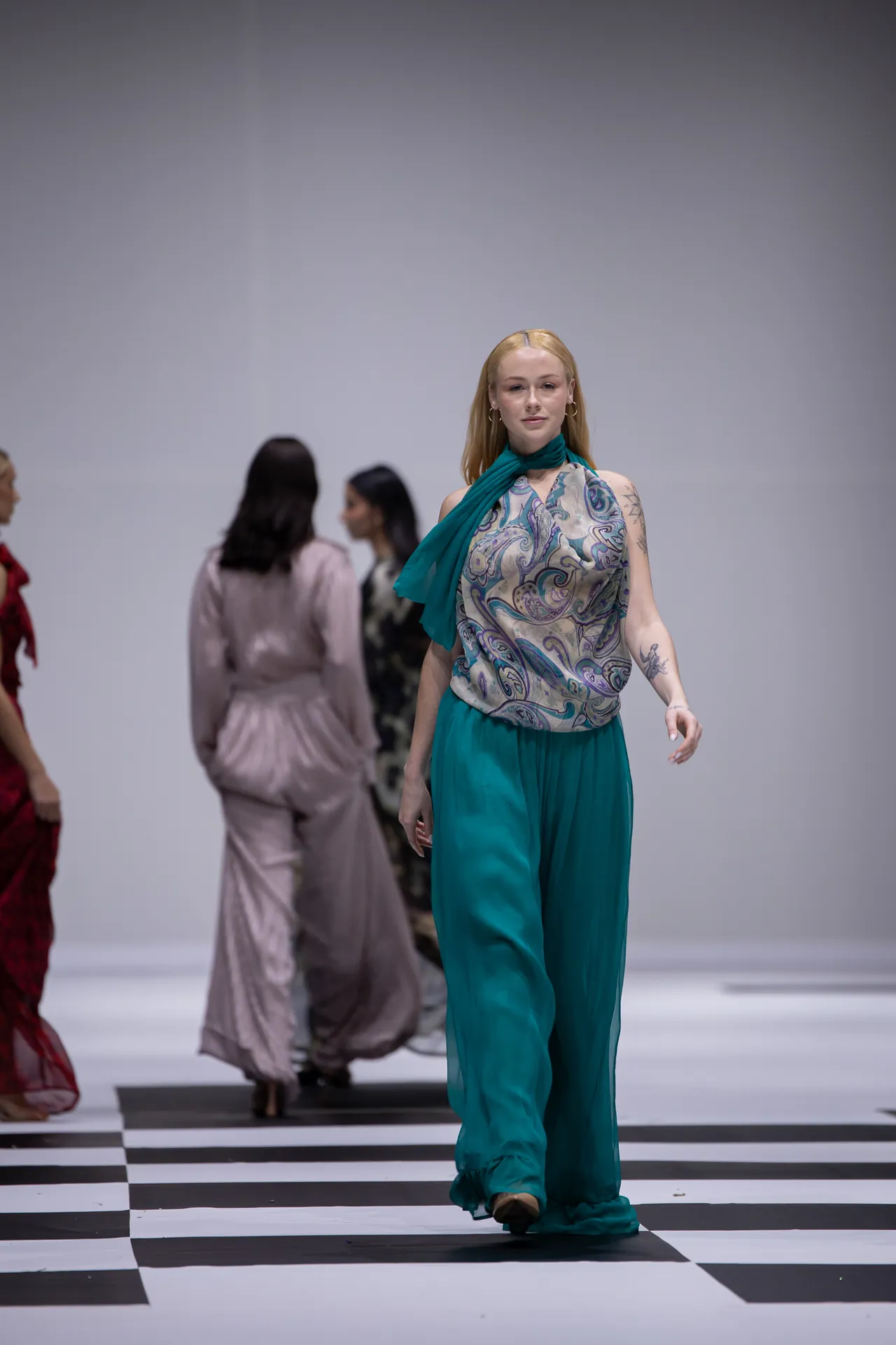 Model walks a fashion runway wearing teal wide-leg pants and a patterned top with a teal scarf.