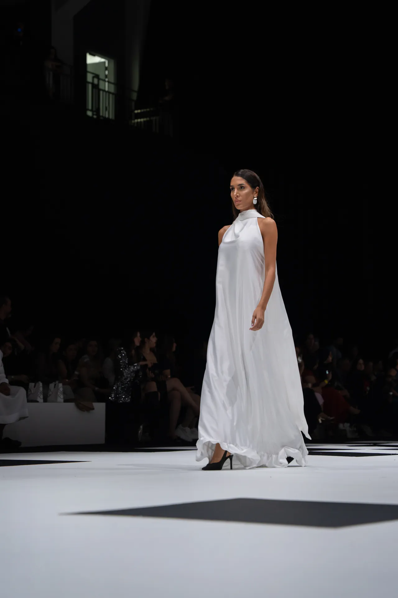 Female model wearing a white, floor-length gown walks the runway at a fashion show on a bright catwalk.