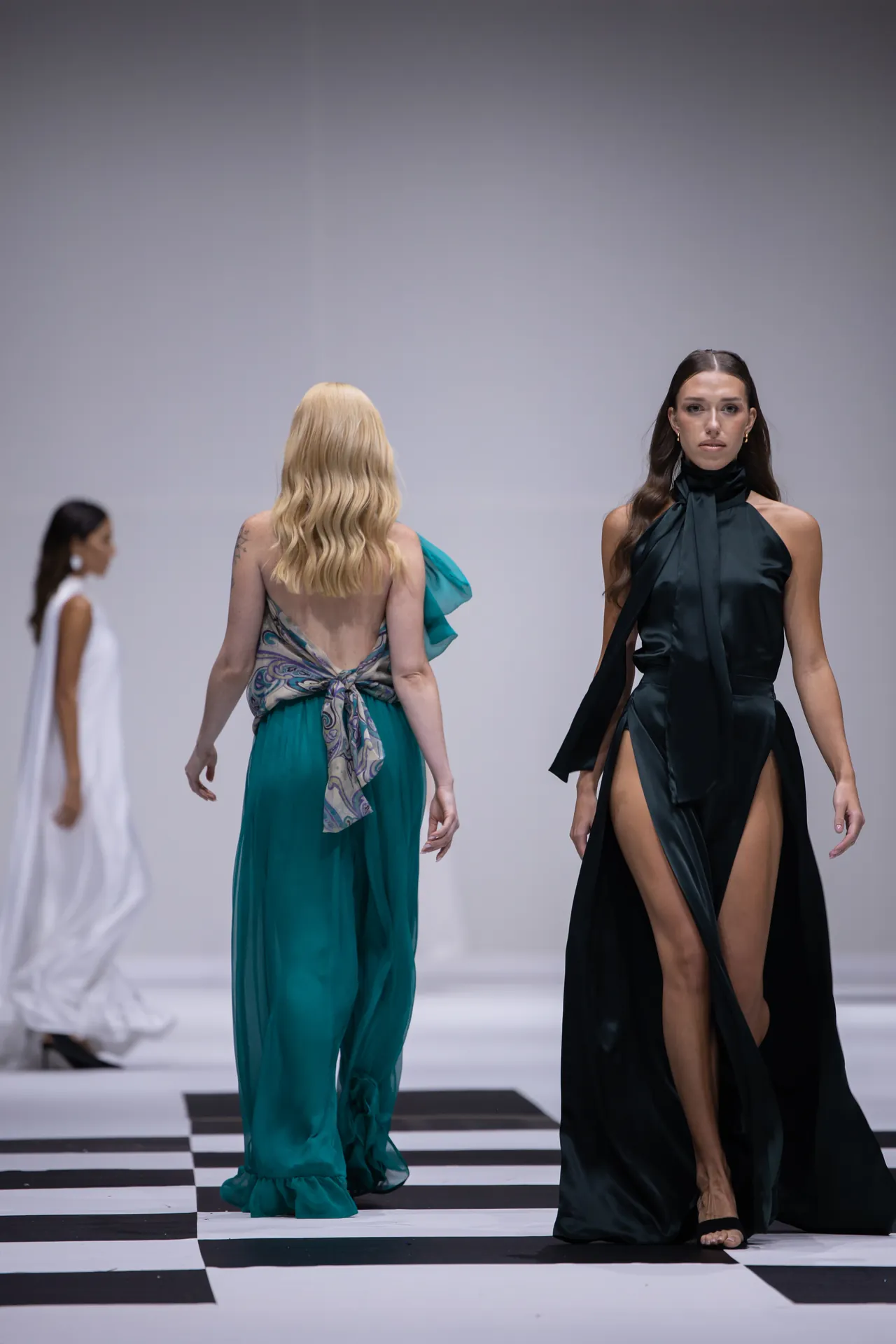 Fashion runway scene with models in formal gowns walking on a black-and-white checkered floor with a plain backdrop.