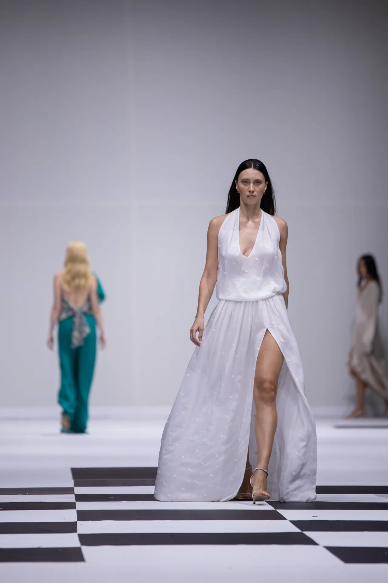 Model walks the runway in a white halter dress with a high slit, on a black-and-white checkered catwalk.