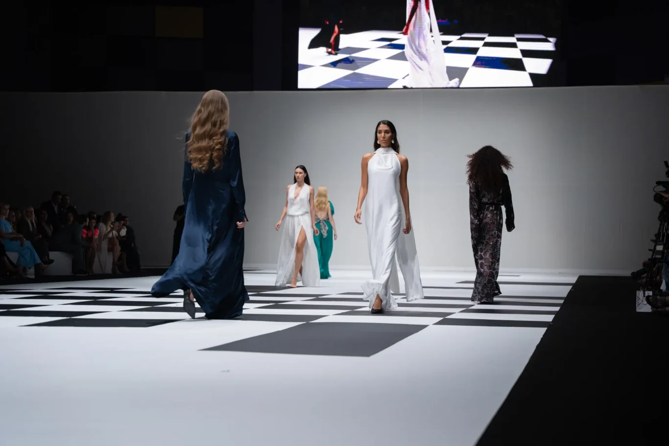 Models walk a fashion show on a black-and-white checkerboard runway in flowing gowns. Textured audience sits along the side.