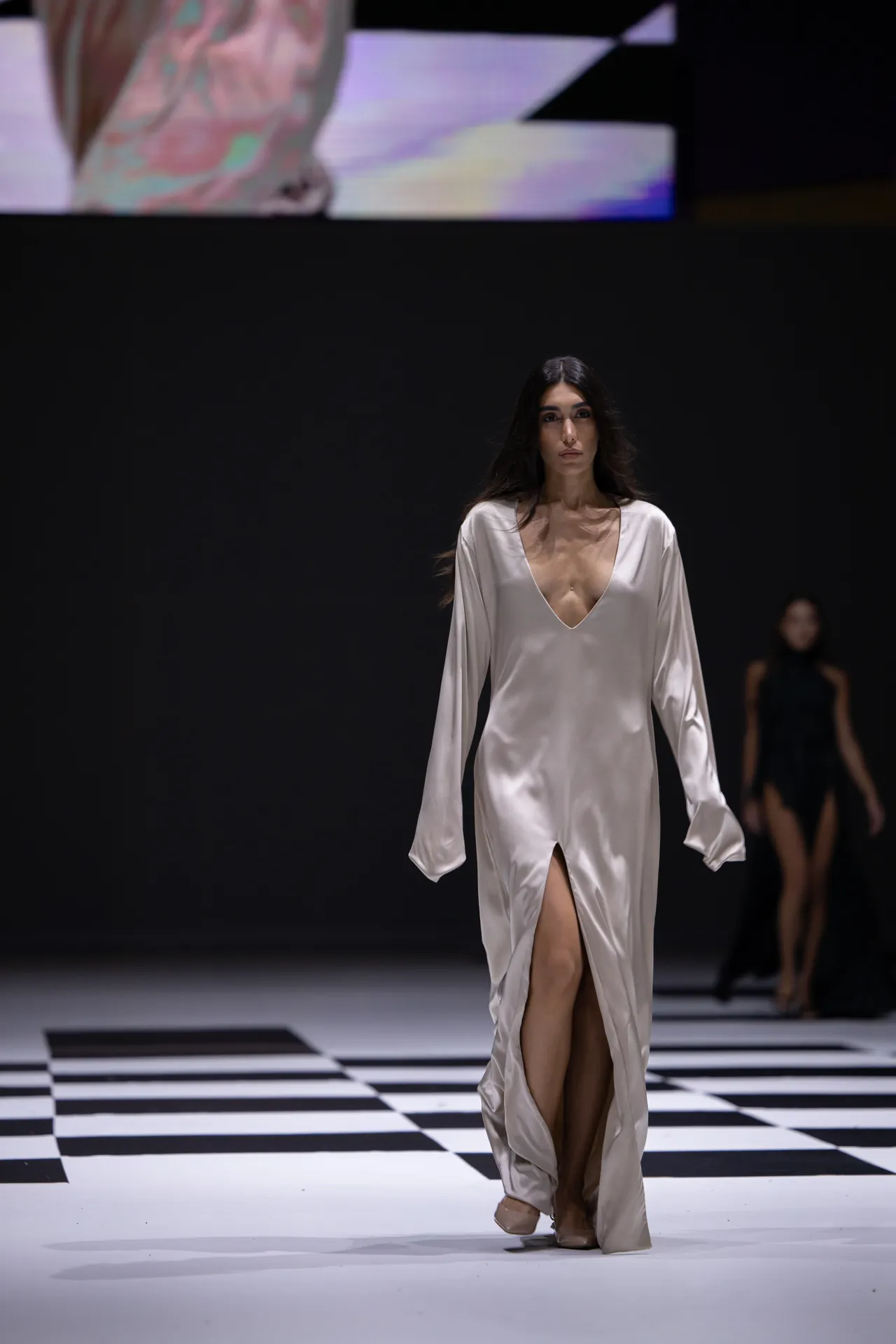 Model walking on a runway in a silky white deep-V gown with a high slit, white floor with black-and-white checkerboard pattern in the foreground.
