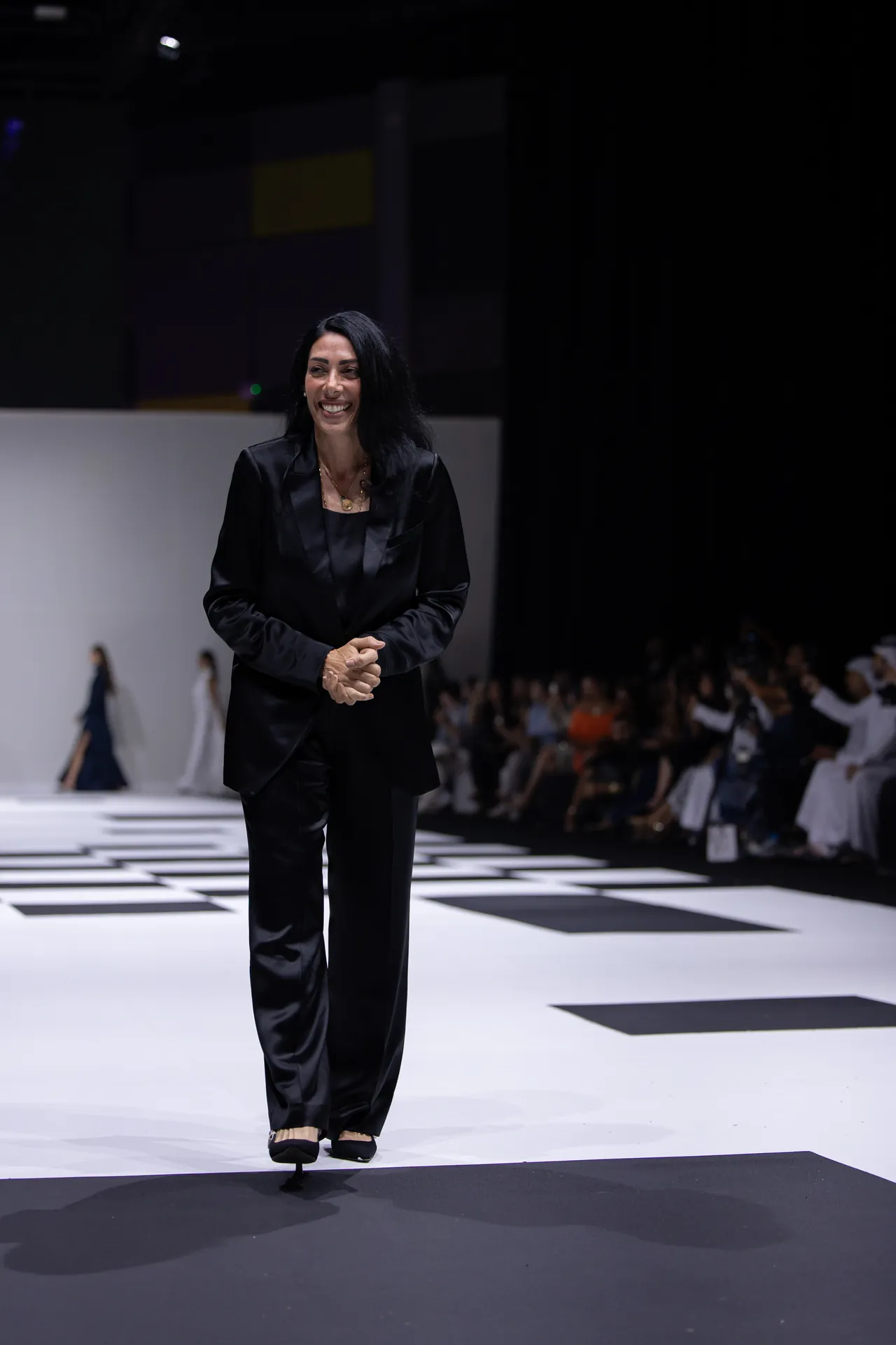 Woman in a black suit walking on a runway, smiling, with a checkered stage floor and an audience in the background.