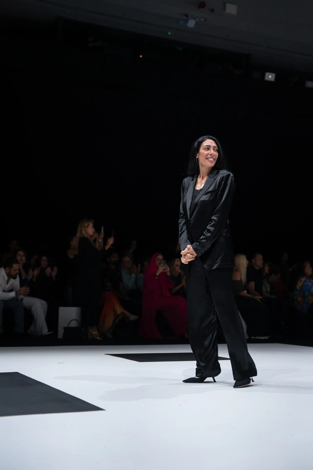 Female model in a black suit walking on a white runway at a fashion show, audience watching in the background.