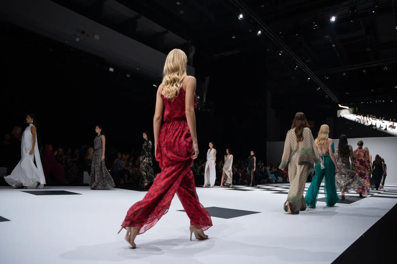 Model in a red patterned dress walks the runway at a fashion show, with other models in colorful outfits following behind.