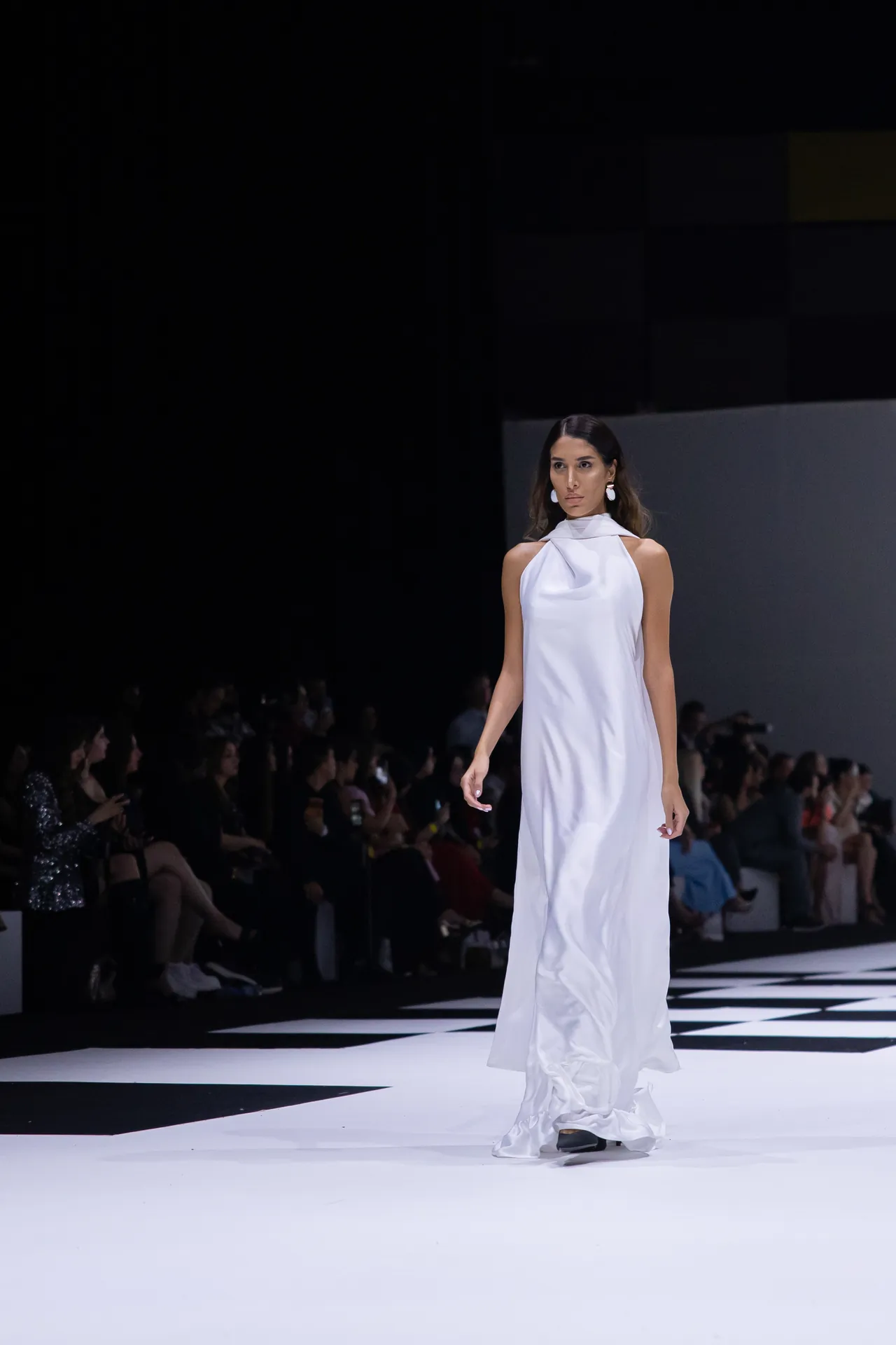 Model wearing a long white satin gown walks the runway as spectators watch from the dimly lit audience area.