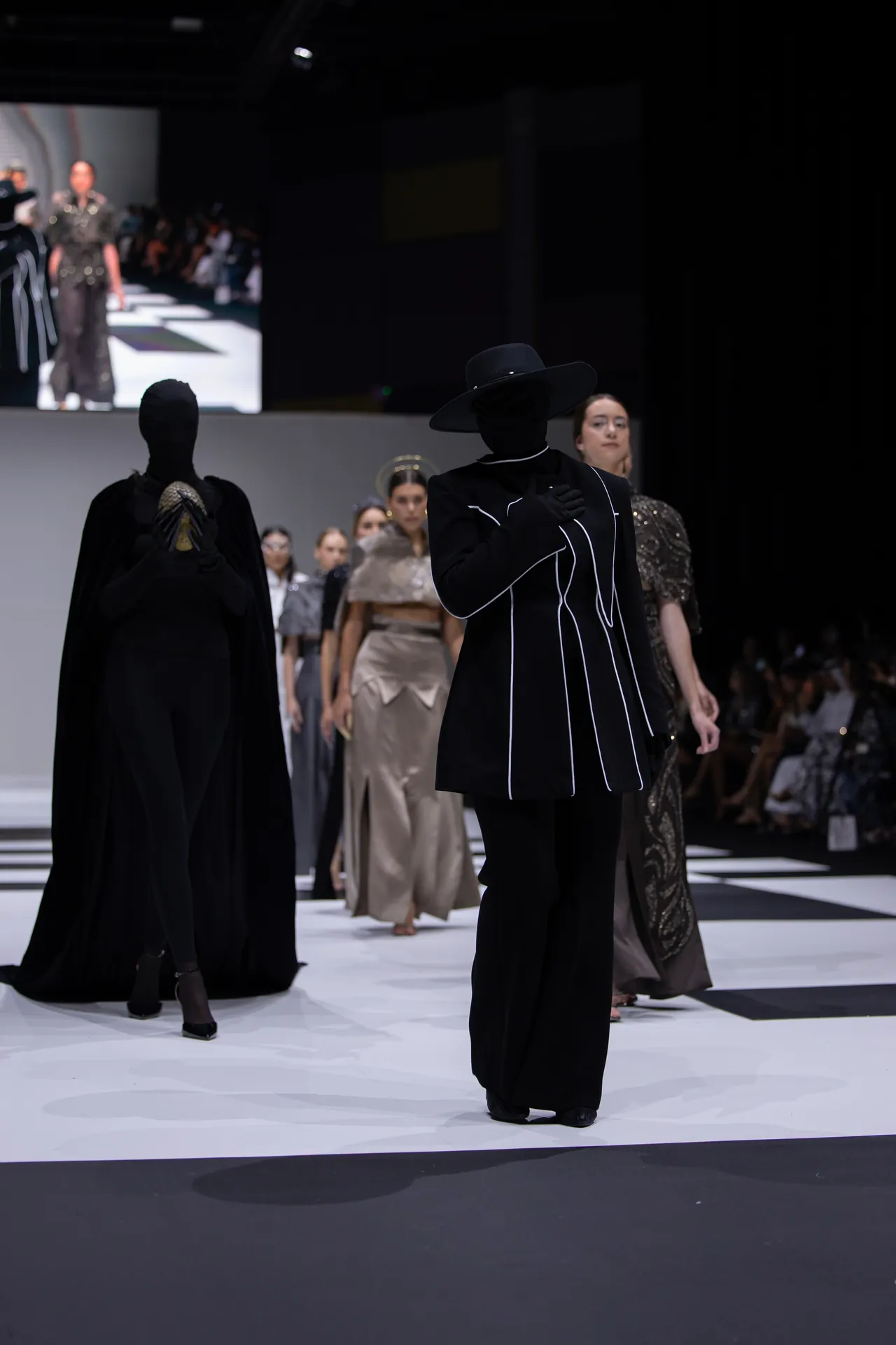 Model in a black coat with white piping and a wide-brim hat walks the runway; blurred audience and other models visible behind him/her.