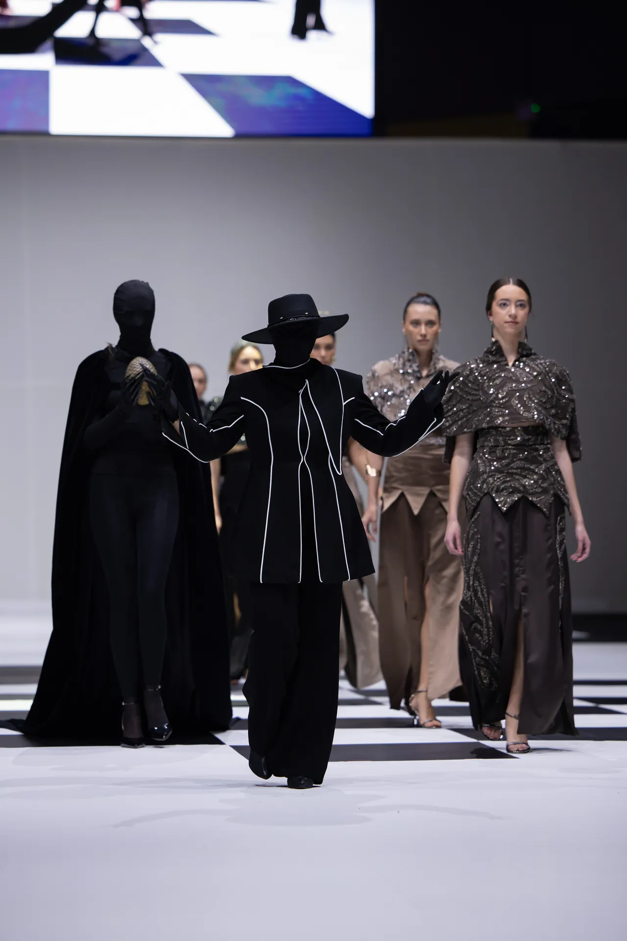 Model in a black outfit with white piping and a wide-brim hat walks the checkerboard runway, followed by other models in ornate gowns.