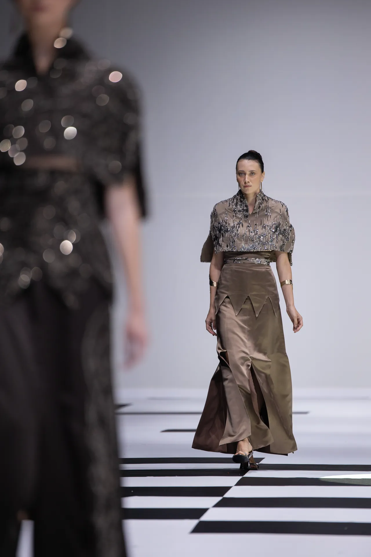 Model on a fashion runway wearing a metallic-beige embellished top with a high-slit satin skirt; a blurred foreground model suggests a catwalk scene and a black-and-white checkered runway.