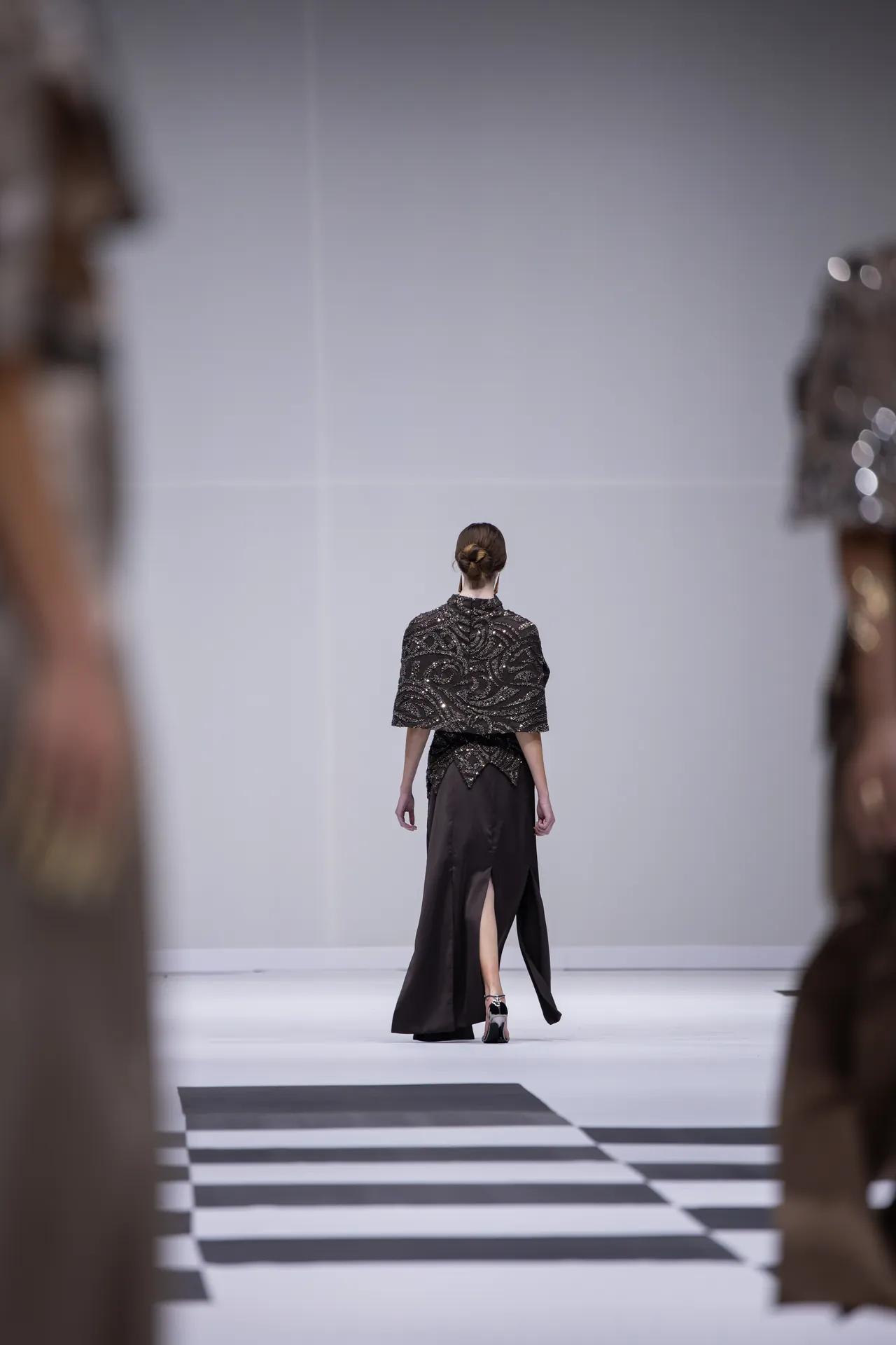 Model walks away down a black-and-white striped runway in a black embellished gown, audience blurred on both sides.