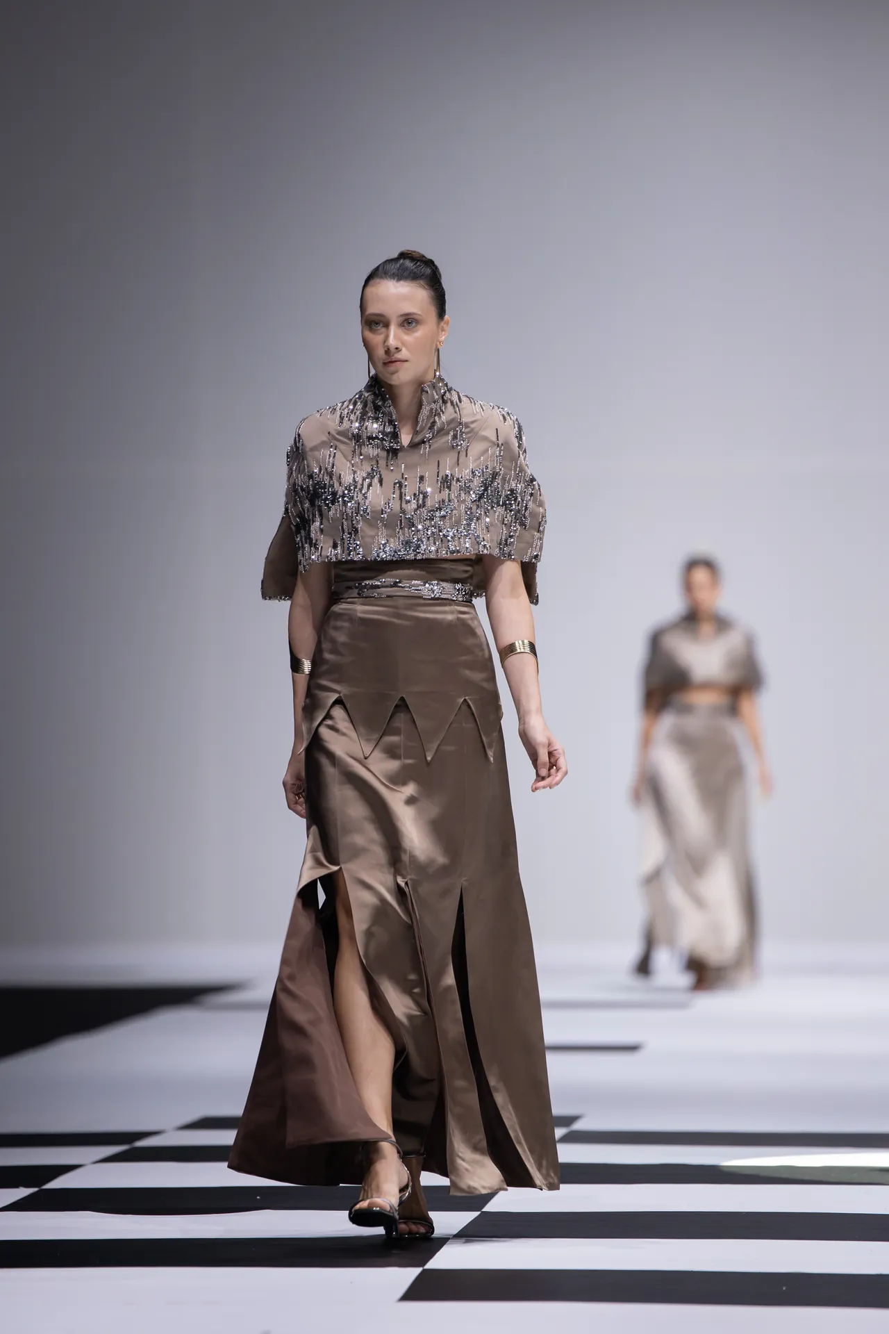 Model walks a fashion show runway in a brown satin dress with a beaded sheer top and metallic details.