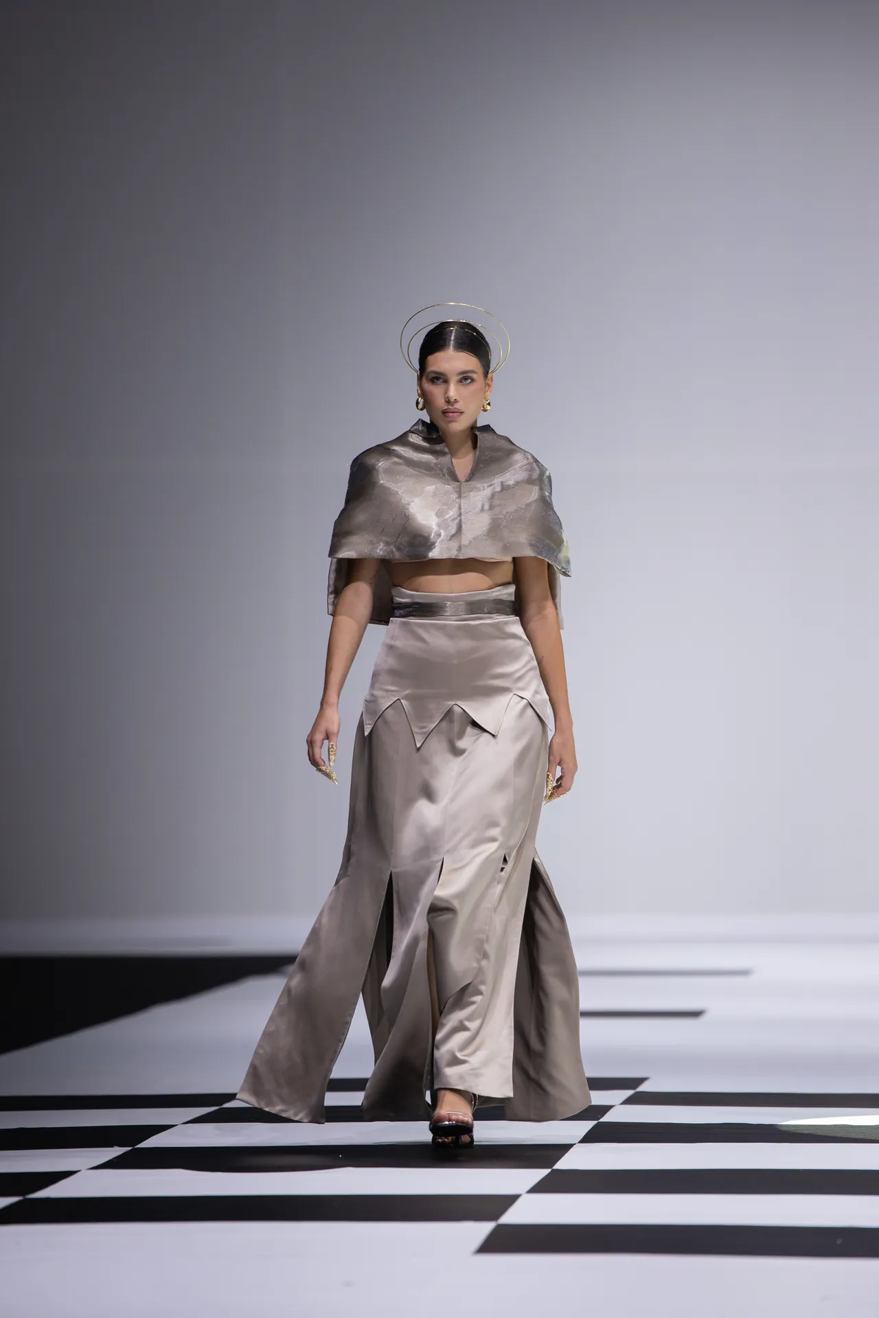 Model walking a runway in a metallic silver two-piece ensemble with a cape and high-slit skirt; black-and-white checkered floor, minimalist backdrop, gold halo headpiece and jewelry