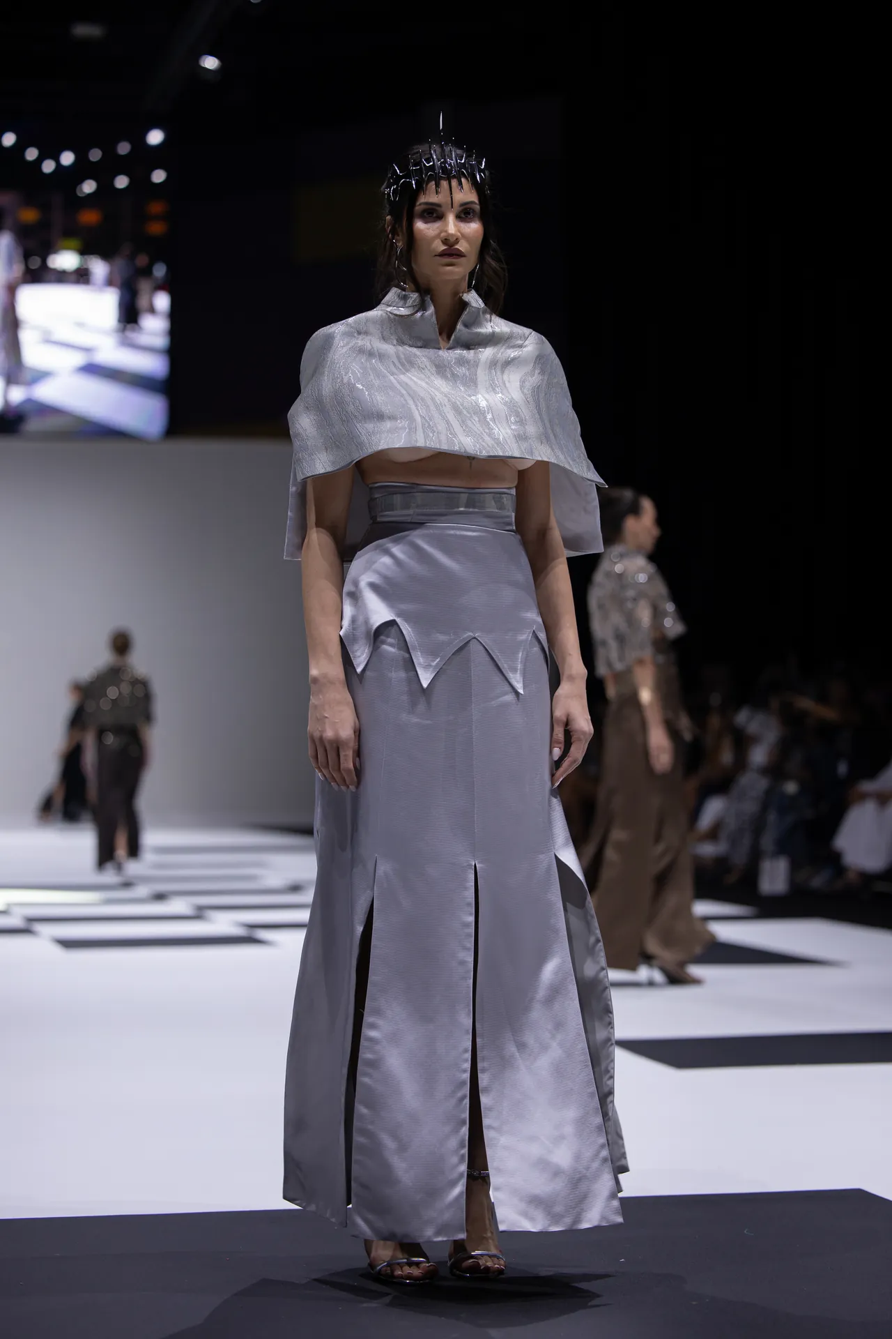 Model on a runway wearing a metallic silver cape top and long skirt with angular panels, paired with a spiked headpiece under studio lighting.