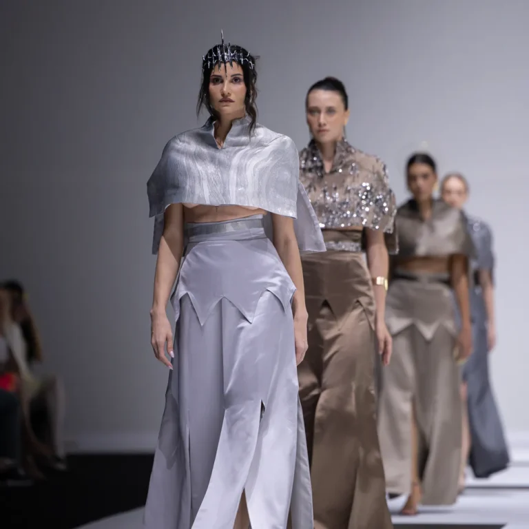 Models walk a fashion runway in metallic silver outfits; the lead model wears a cropped cape and angular skirt with a headpiece of spikes.