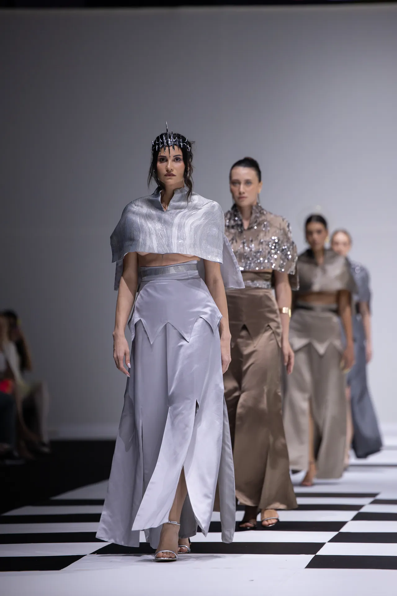 Models walk a fashion runway in metallic silver outfits; the lead model wears a cropped cape and angular skirt with a headpiece of spikes.