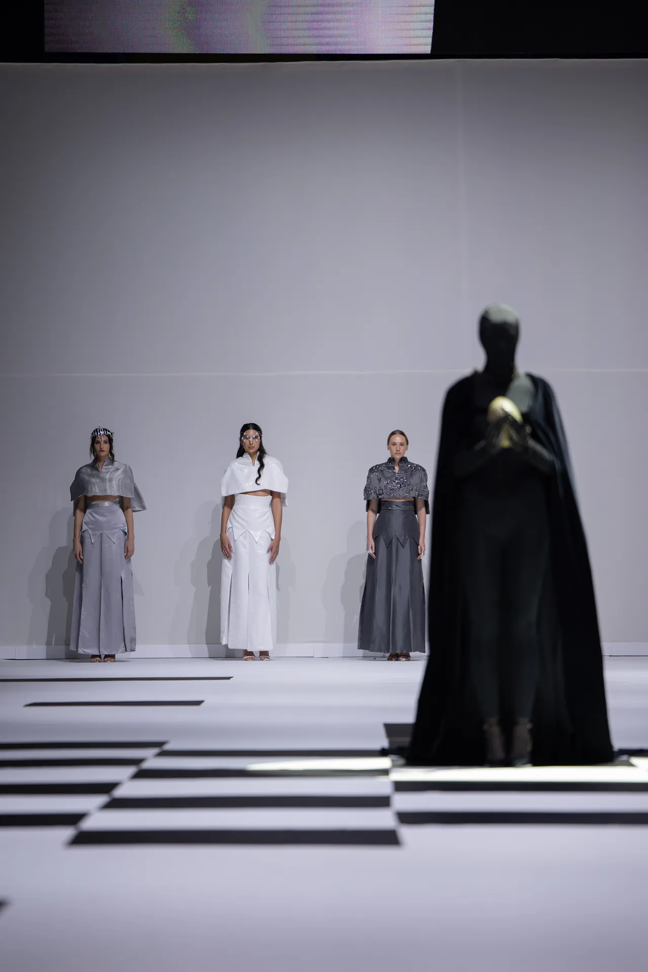 Four models in metallic gray and white gowns stand on a white runway, with a large dark figure in the foreground right.