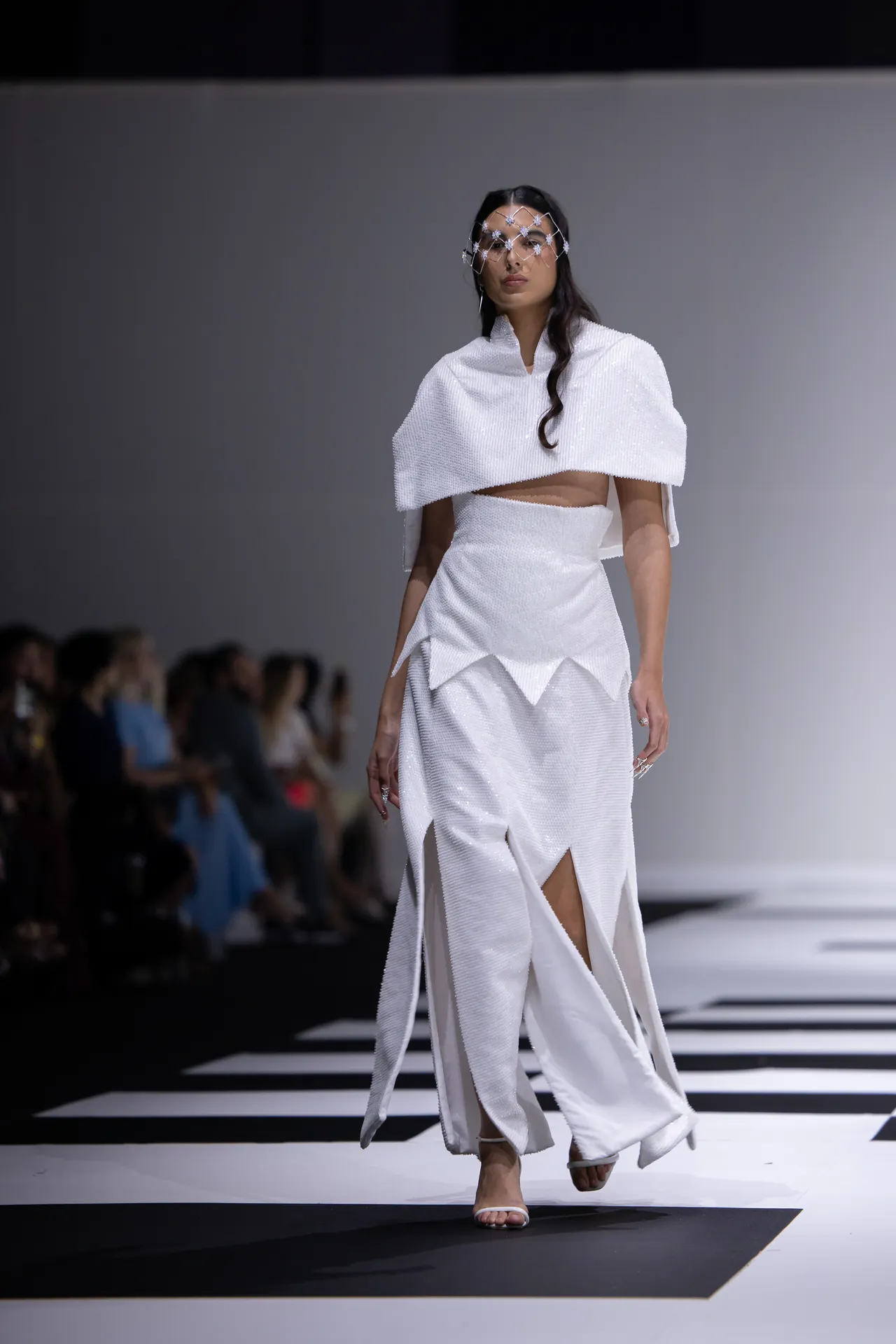 Female model walks a white textured two-piece outfit on a fashion runway, wearing a decorative crystal headpiece and open-toed heels.