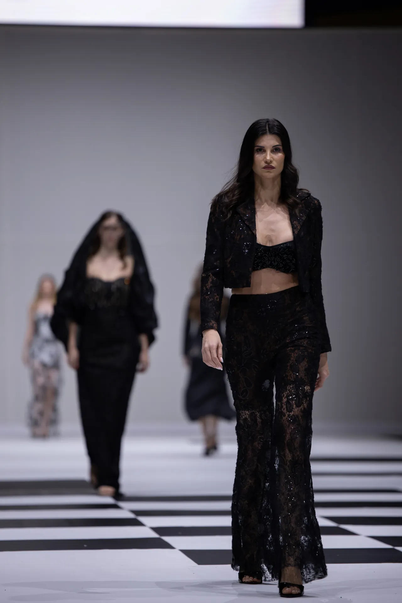 Model in a black lace two-piece outfit walking a fashion runway with a black-and-white checkered floor in the foreground.