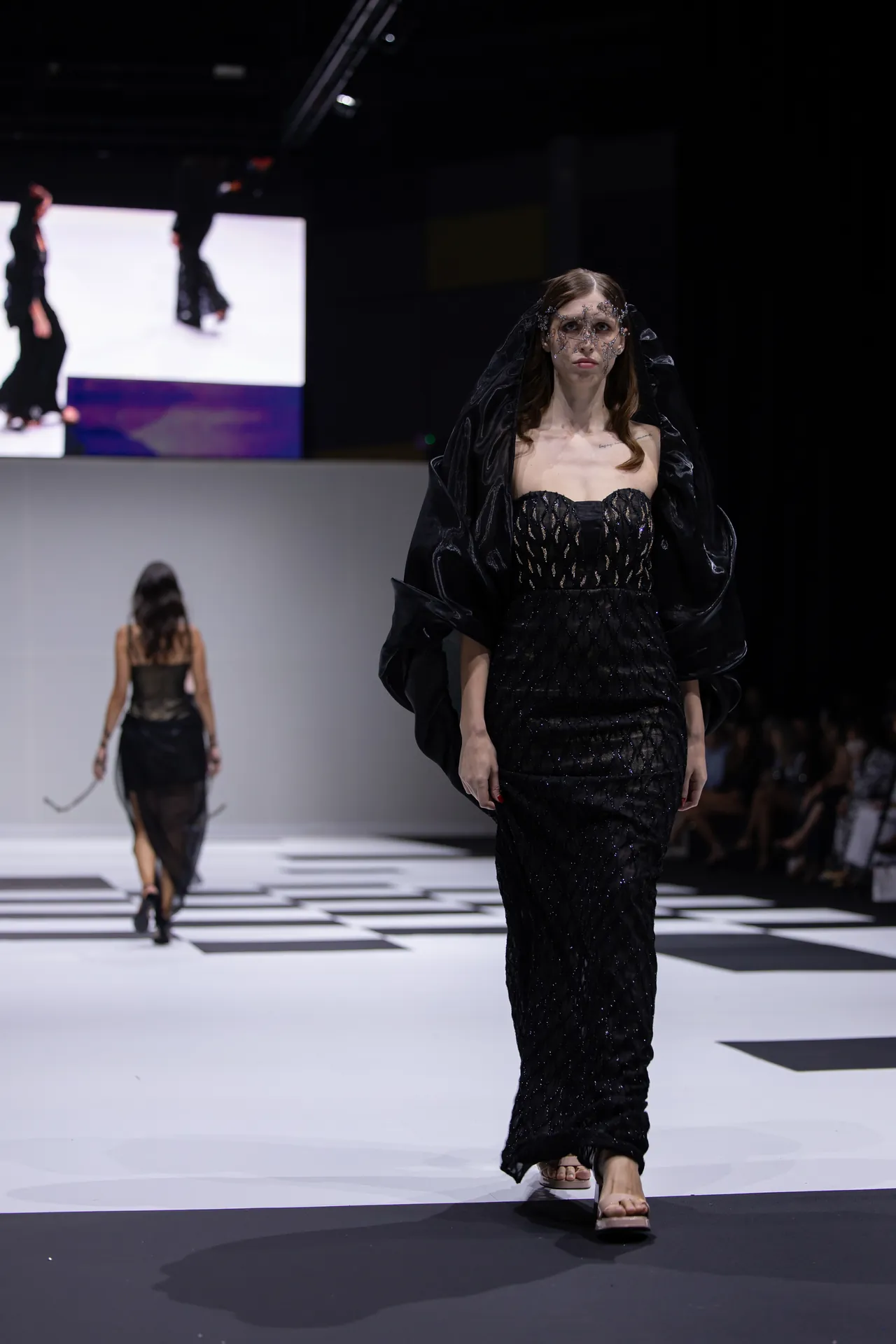 Model walks a black embellished gown with a sheer veil on a black-and-white checkered runway at a fashion show, with another model and screens in the background.