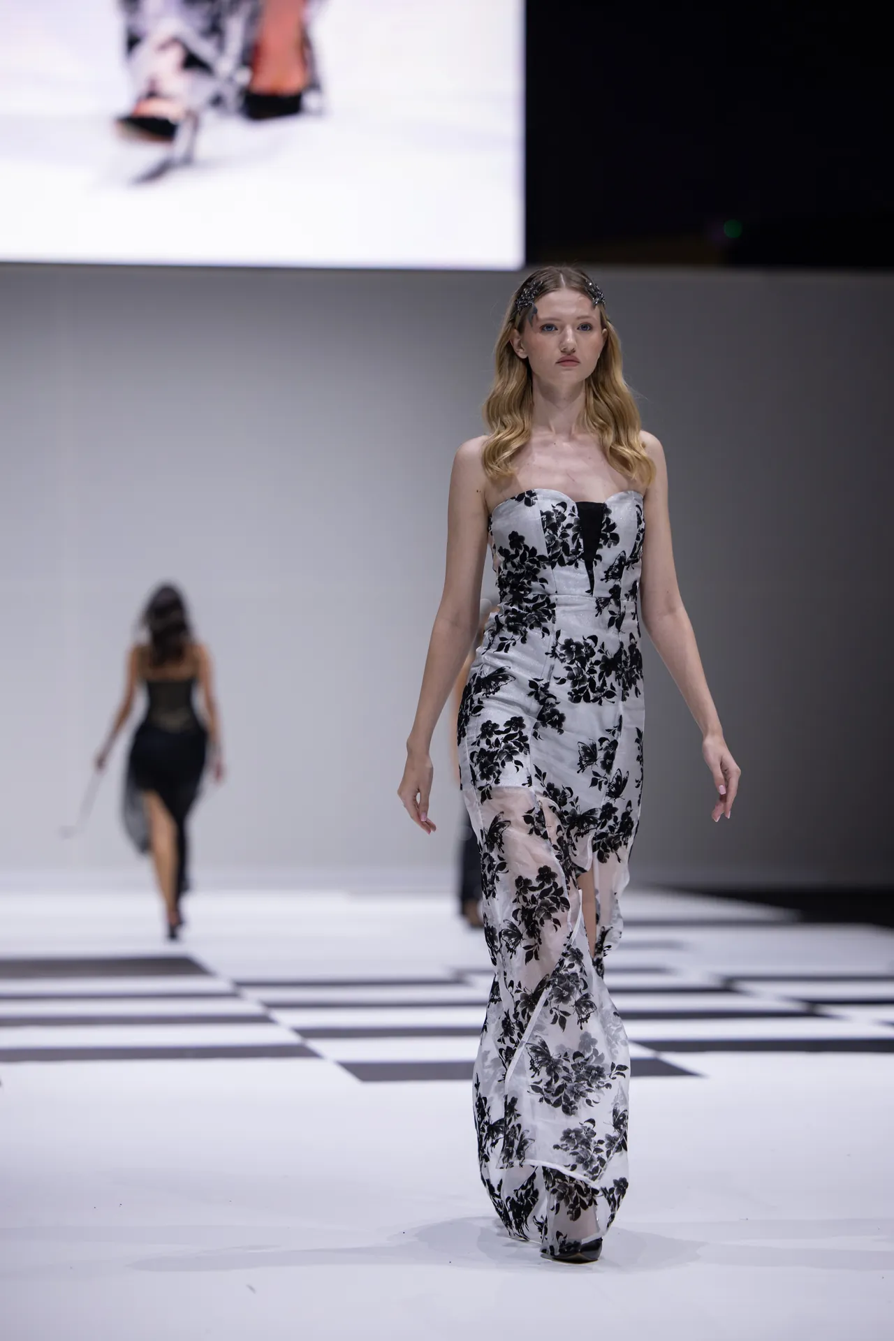 Model walks a white, black-floral strapless gown down the runway with confidence.