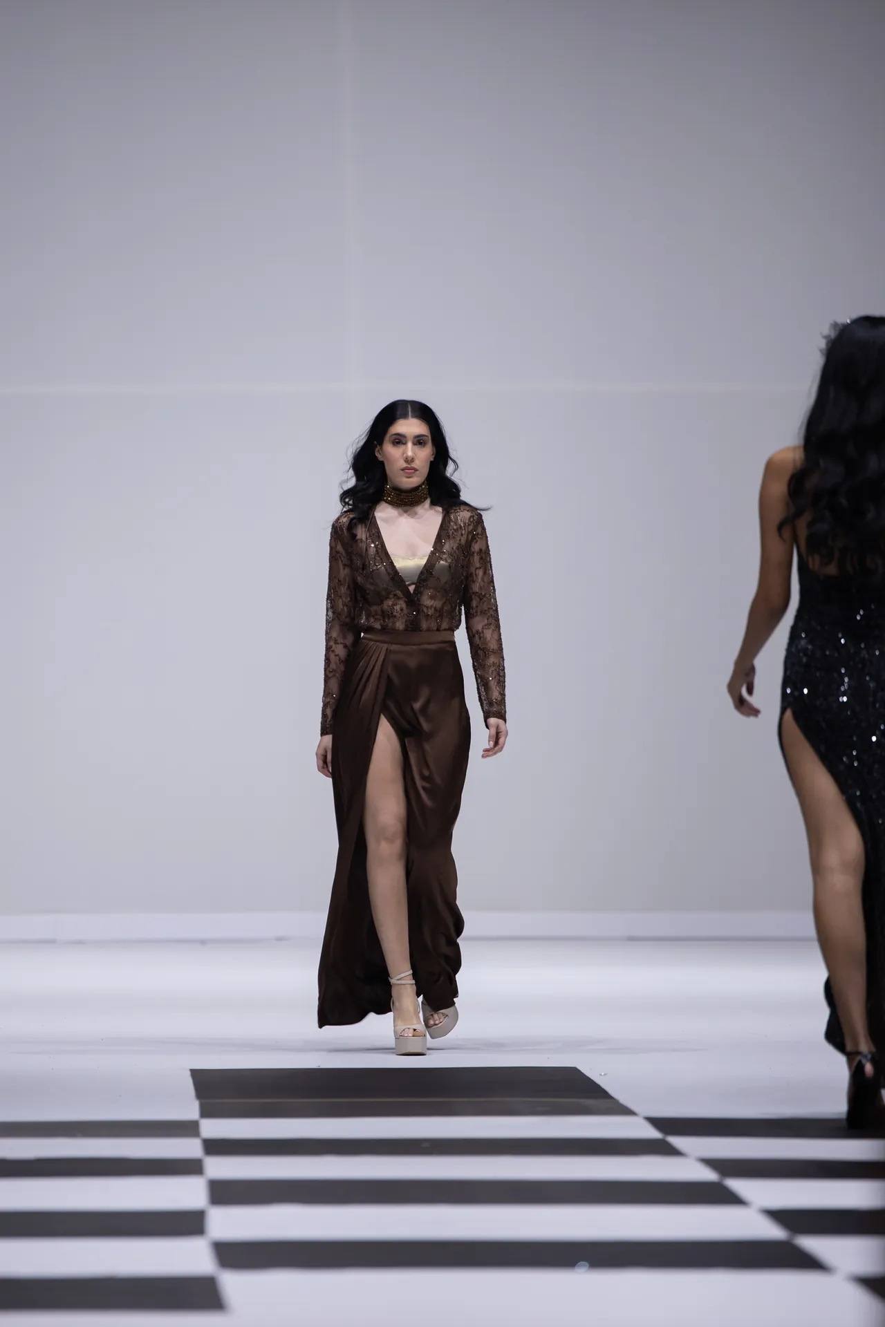 Model walking a fashion runway in a brown lace long-sleeve top and high-slit satin skirt on a white catwalk with another model visible to the side.