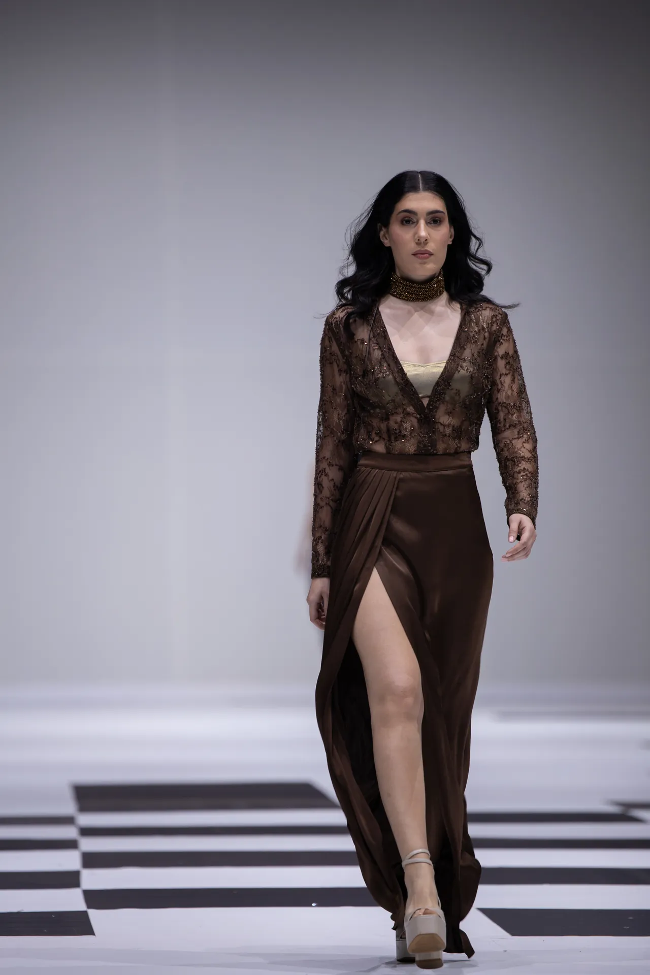 Female model walking a runway in a brown lace blouse and satin wrap skirt with a gold choker necklace and platform sandals.